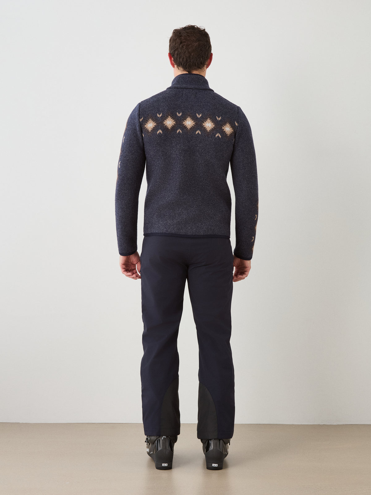 A man stands facing away, wearing the We Norwegians Hovden Jacket Men—crafted from navy boiled merino wool and featuring a geometric pattern on the back inspired by Norwegian Sámi design—paired with dark pants and black ski boots, against a plain light background.