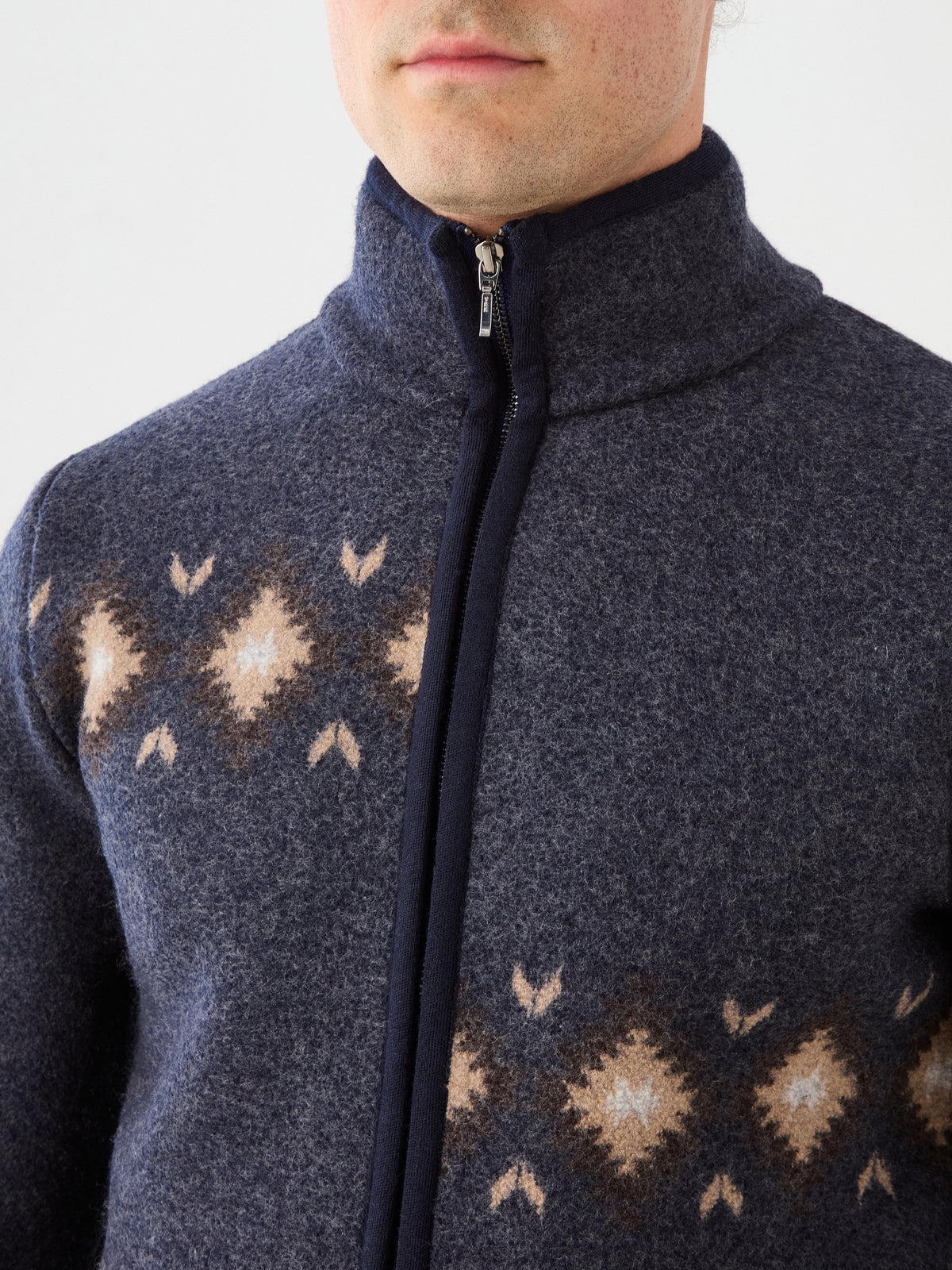 A person wearing the We Norwegians Hovden Jacket Men—a zip-up blue sweater featuring a tan and white diamond pattern across the chest reminiscent of Norwegian Sámi designs. The photo is cropped to show only the lower face and upper body.