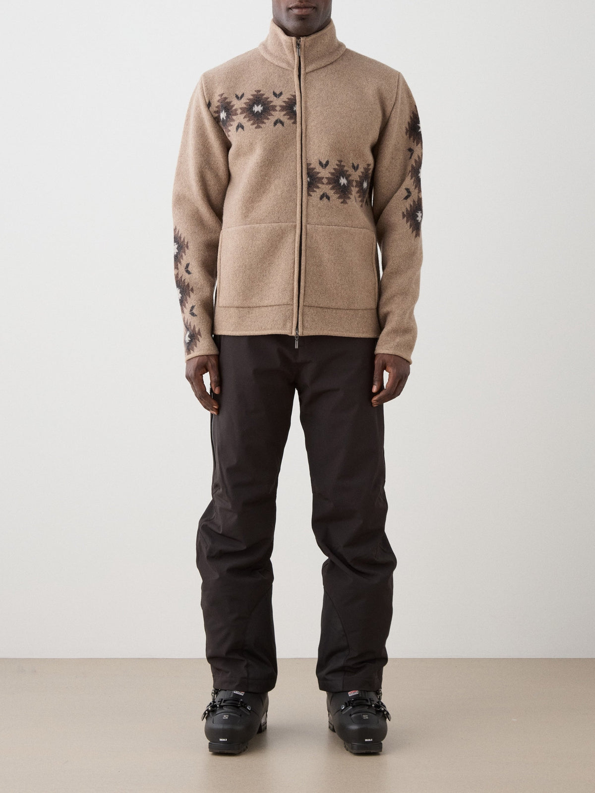 A person wearing a beige, full-zip We Norwegians Hovden Jacket Men made of boiled merino wool with a dark geometric pattern on the chest and sleeves, paired with black pants and ski boots.