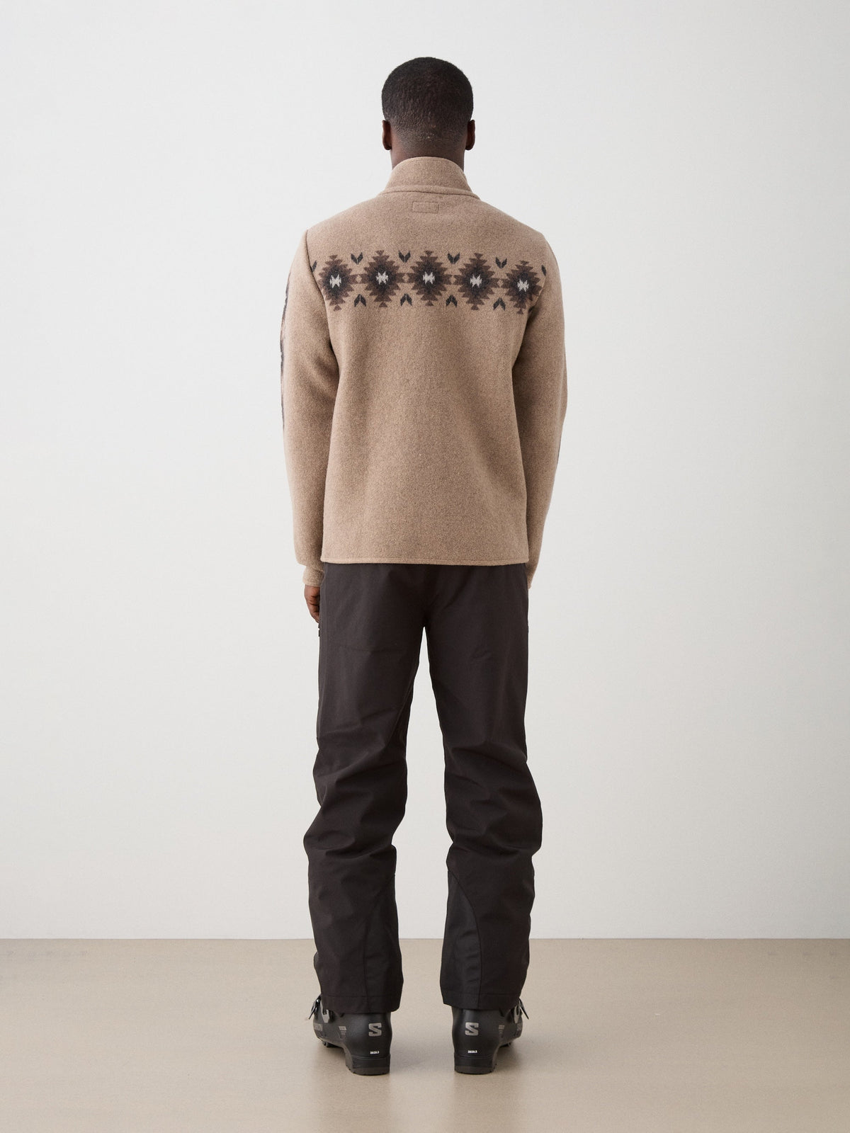 Back view of a person wearing a beige, midweight wool Hovden Jacket Men with a dark geometric pattern across the upper back, paired with dark pants and ski boots.