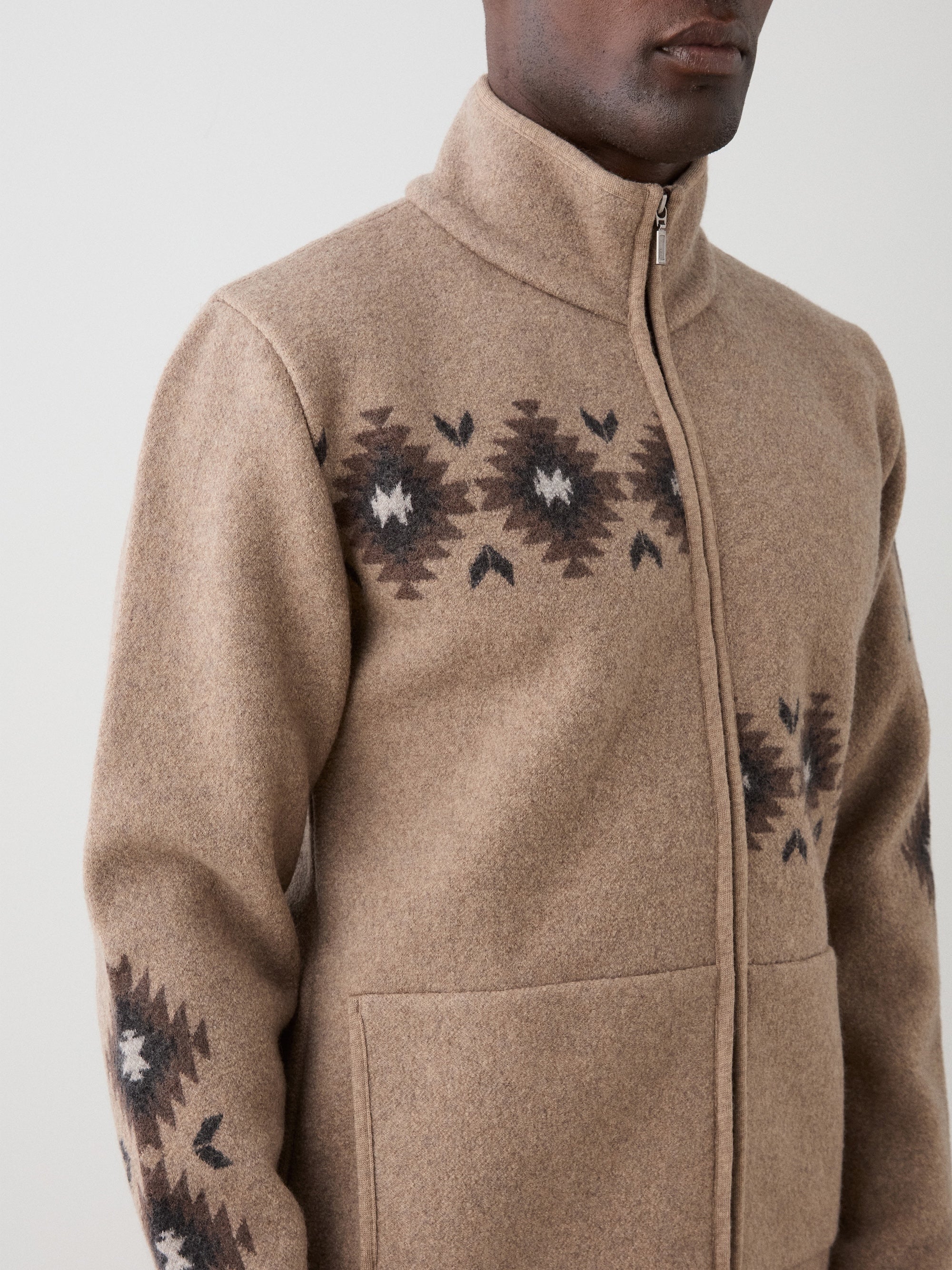A person wears the Hovden Jacket Men by We Norwegians—tan boiled Merino wool with black and white Sámi-inspired geometric chest and sleeve patterns—featuring a high collar, midweight warmth, and two front pockets, against a plain light background.