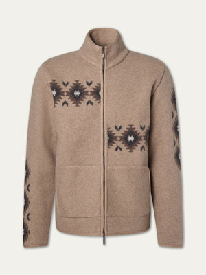 The We Norwegians Hovden Jacket Men is a tan, Sámi-inspired zip-up boiled Merino wool jacket with a high collar, two front pockets, and black geometric patterns with white accents on the chest, sleeves, and sides.