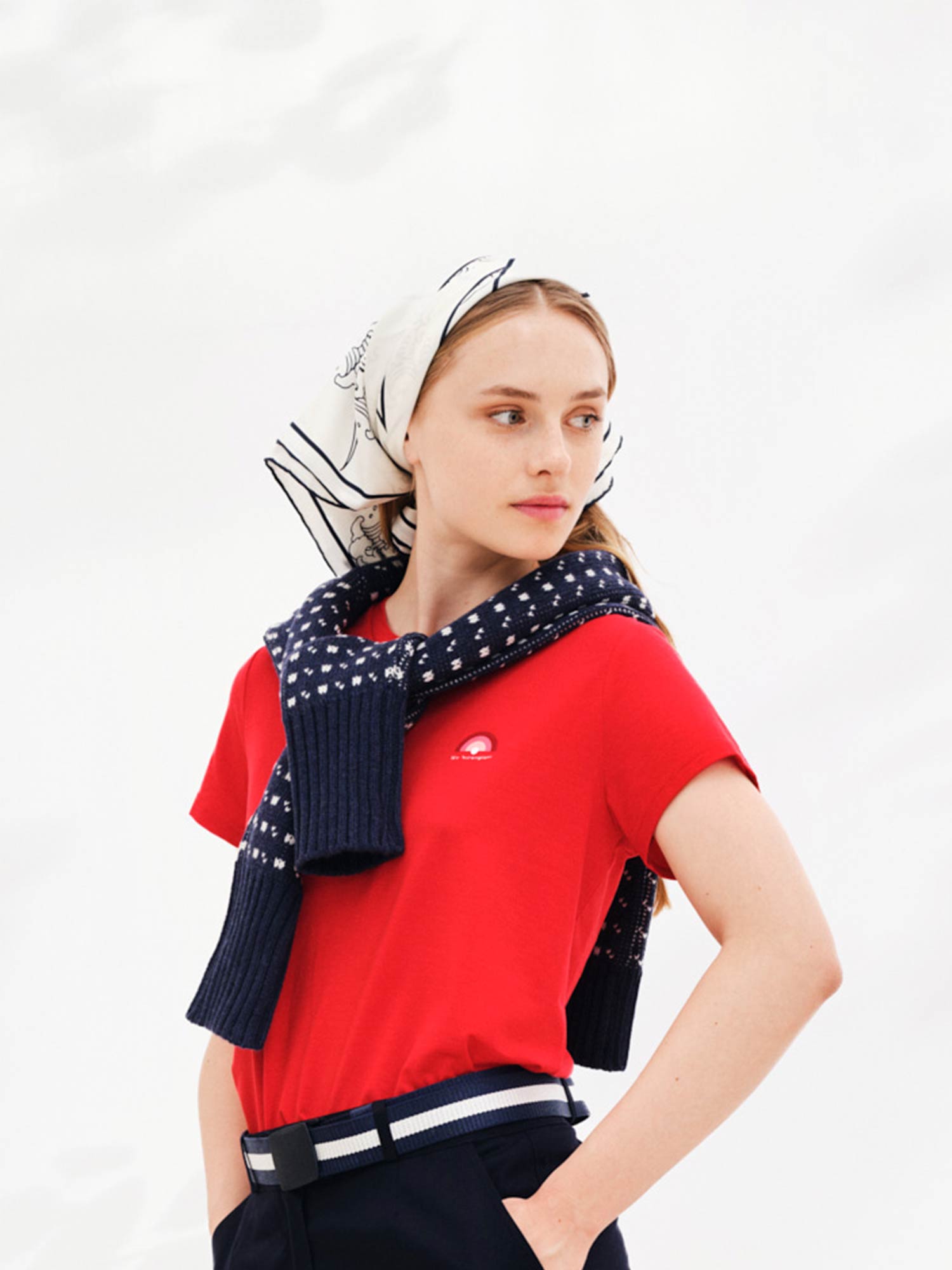 A young woman stands against a plain white background, wearing a red t-shirt, dark pants with a white belt, and the Wavy Silk Scarf White from We Norwegians draped over her shoulders. She also has a white headscarf with black designs. She looks slightly to the side, evoking the elegance of Ukiyo-e art.