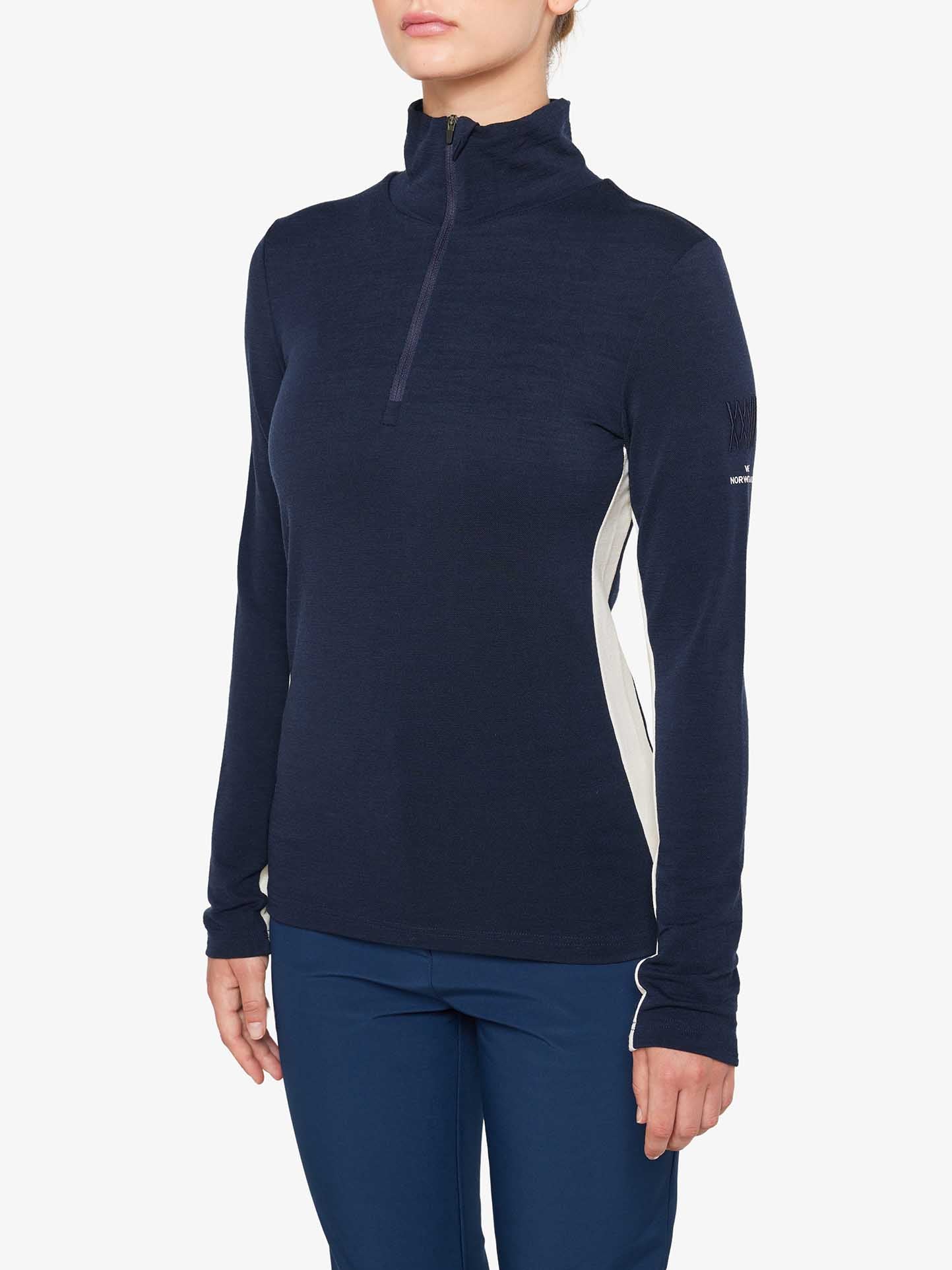 A person is wearing a navy blue, long-sleeved "Voss 1/4-Zip Women" pullover by *We Norwegians,* featuring white color block panels and matching navy blue pants. Their hands are by their sides, set against a plain white background. Only part of the face is visible, with the focus on the Merino wool clothing.