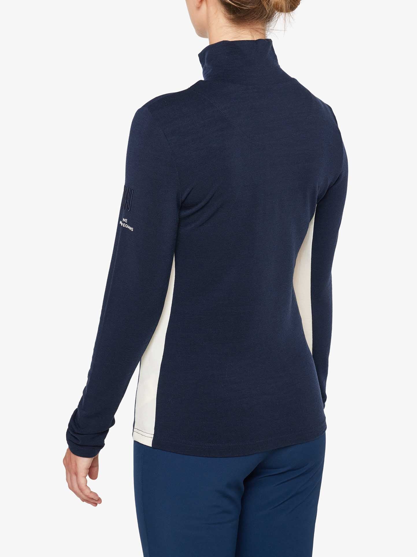 A person with blonde hair is seen from the back, wearing a navy blue and white Voss 1/4-Zip Women athletic top by We Norwegians and blue pants. The Merino wool top has a high collar, is fitted, and features color block panels on the sides. The person stands against a plain, light background.