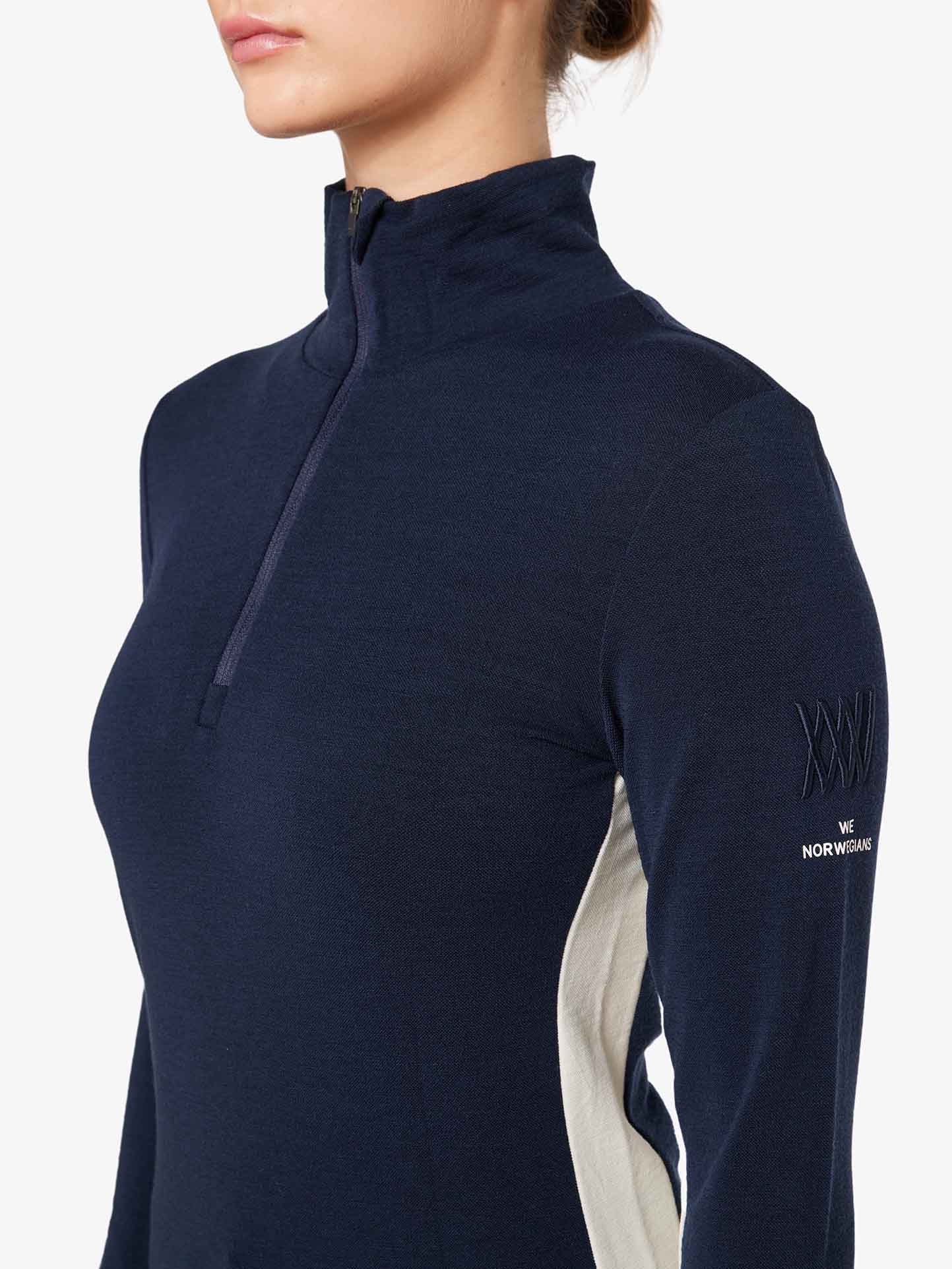 A person dressed in the Voss 1/4-Zip Women pullover by We Norwegians, featuring a dark blue Merino wool fabric with a high collar and light-colored side panels. The sleeve is embroidered with "We Norwegians Norway." The background is plain white, and the photo showcases only their clothing and upper bust, with their face out of frame.