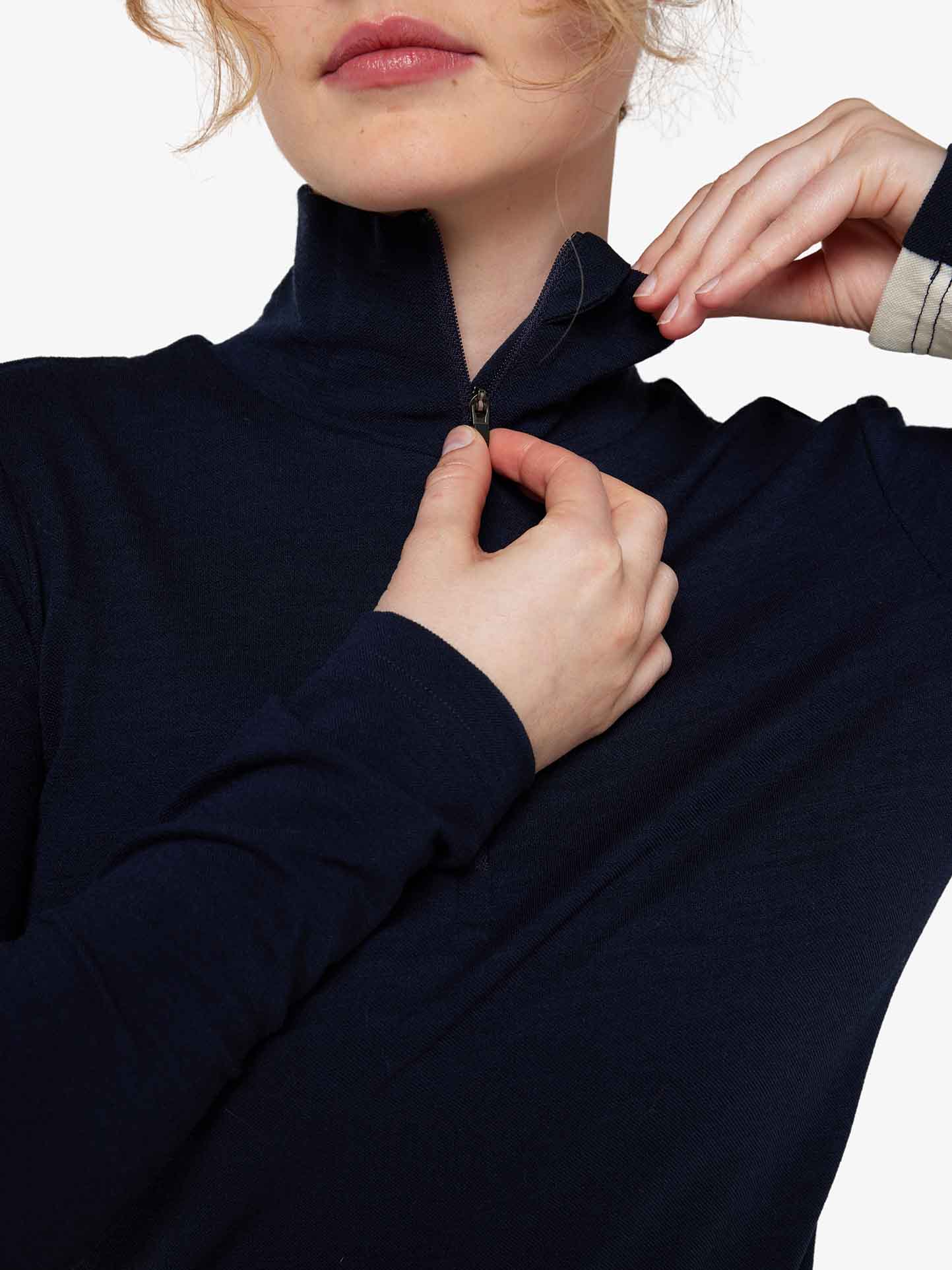 A person wearing a long-sleeve dark blue Voss 1/4-Zip Women by We Norwegians, featuring color block panels, is partially unzipping the high collar. The upper part of their face is not visible, and their hands are positioned near the zipper and the collar.