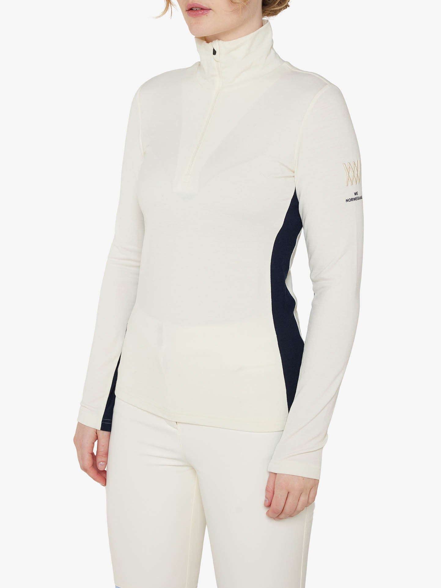 A person is shown wearing We Norwegians' Voss 1/4-Zip Women long-sleeved athletic top in white, which features a high collar and a partial front zipper. The Merino wool top includes dark side color block panels and a small logo on the left sleeve. The person is also wearing matching white athletic pants.