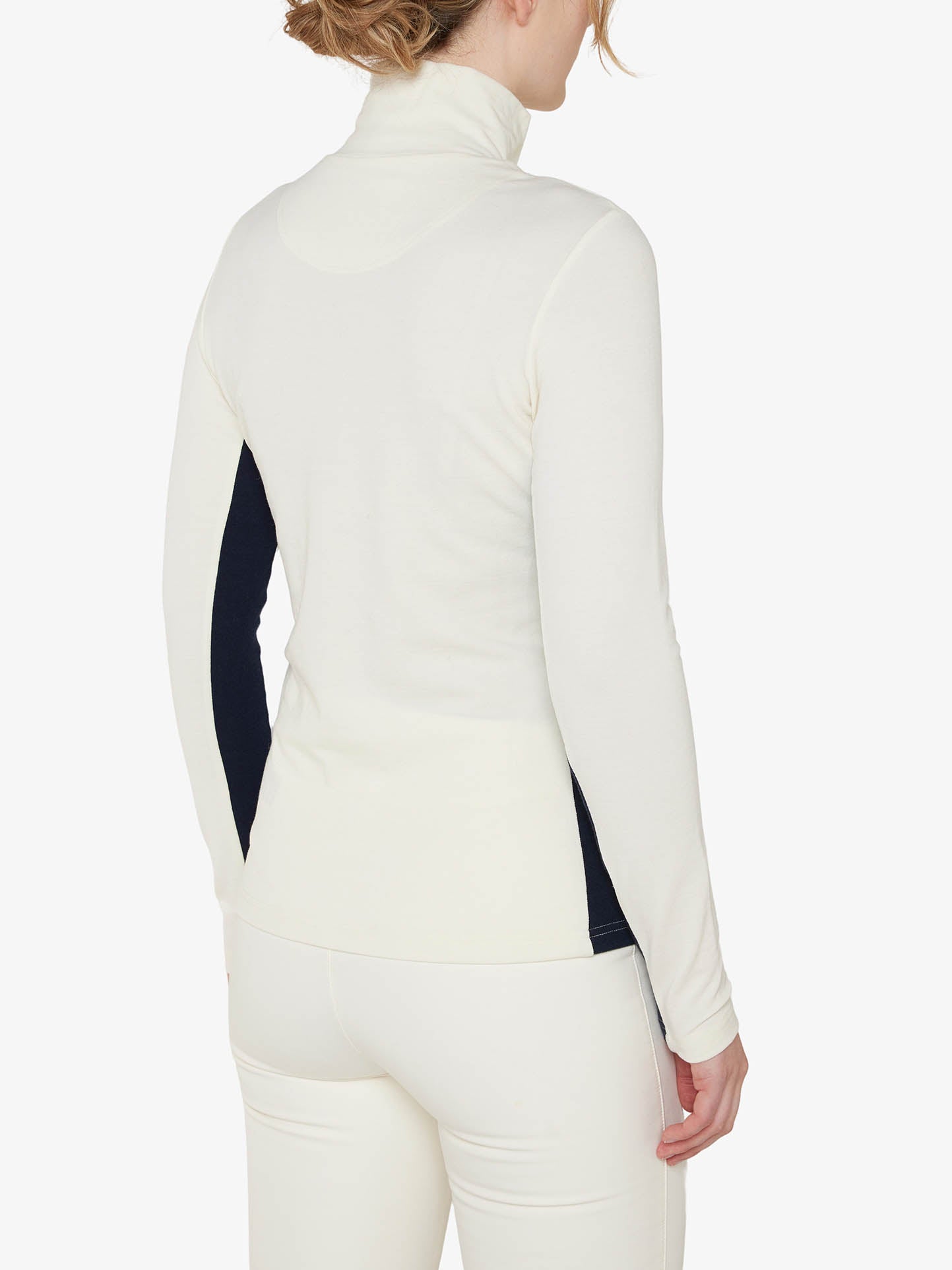 A person wearing a white Voss 1/4-Zip Women shirt by We Norwegians and white pants is shown from the back. The shirt features black color block panels on the sides. The individual has light brown hair tied up and stands against a plain white background.