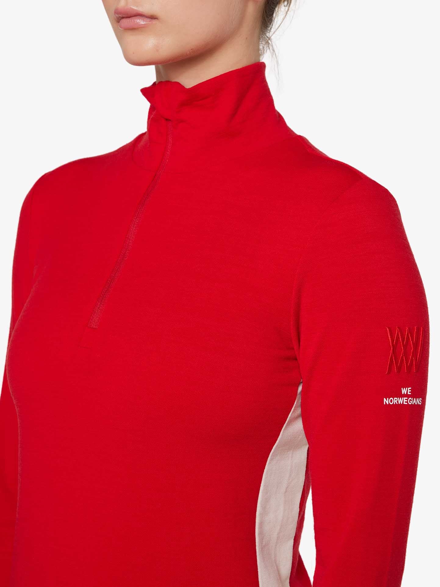 A person wearing the Voss 1/4-Zip Women by We Norwegians, a red long-sleeve top with a zippered collar. Made of Merino wool, the top features white color block panels and text on the sleeve that reads "WE NORWEGIANS." The image is cropped to show only the person's torso and part of their face.