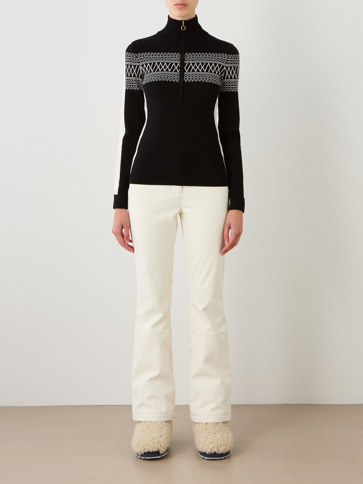 A person wearing the We Norwegians Signature 1/4-Zip Women sweater in a black and white Setesdal pattern, crafted from soft Merino wool, is paired with white pants and fuzzy beige boots while standing against a plain background.