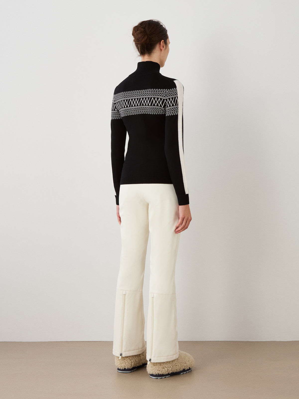 A person with hair in a bun wears the We Norwegians Signature 1/4-Zip Women sweater in black and white with a Setesdal pattern, paired with white pants and fluffy beige boots, standing indoors facing a plain white wall.