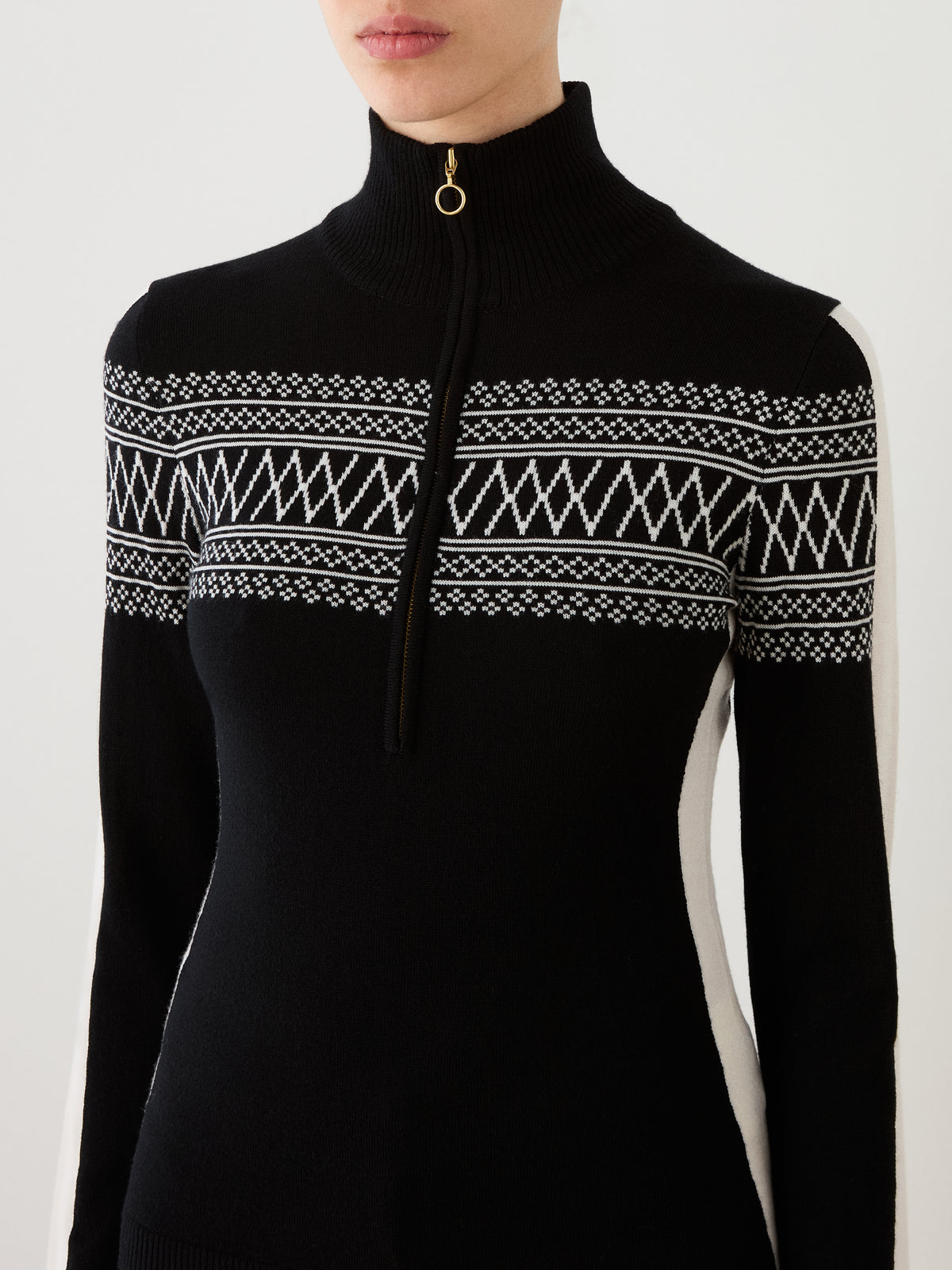 A woman wears the We Norwegians Signature 1/4-Zip Women sweater, a black and white knit featuring geometric patterns inspired by the classic Setesdal design, high collar, distinctive ring-pull quarter-zip, and long sleeves. Only her upper body is visible against a plain background.