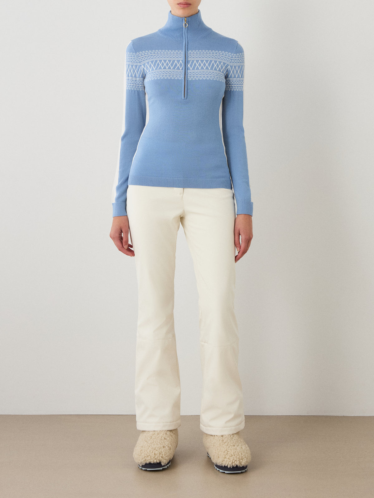 A person wearing the We Norwegians Signature 1/4-Zip Women sweater in light blue, which features a subtle Setesdal pattern, pairs it with off-white pants and beige fluffy shoes while standing against a plain white wall on a light brown floor.