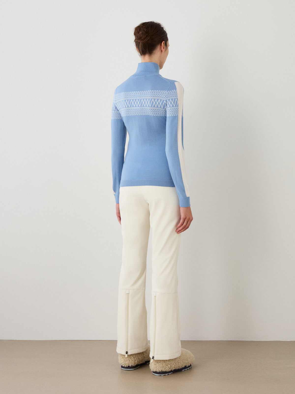A woman stands facing away, wearing the We Norwegians Signature 1/4-Zip Women sweater in light blue with white Setesdal patterns, styled with white pants and beige boots, set against a plain white wall and tan floor.