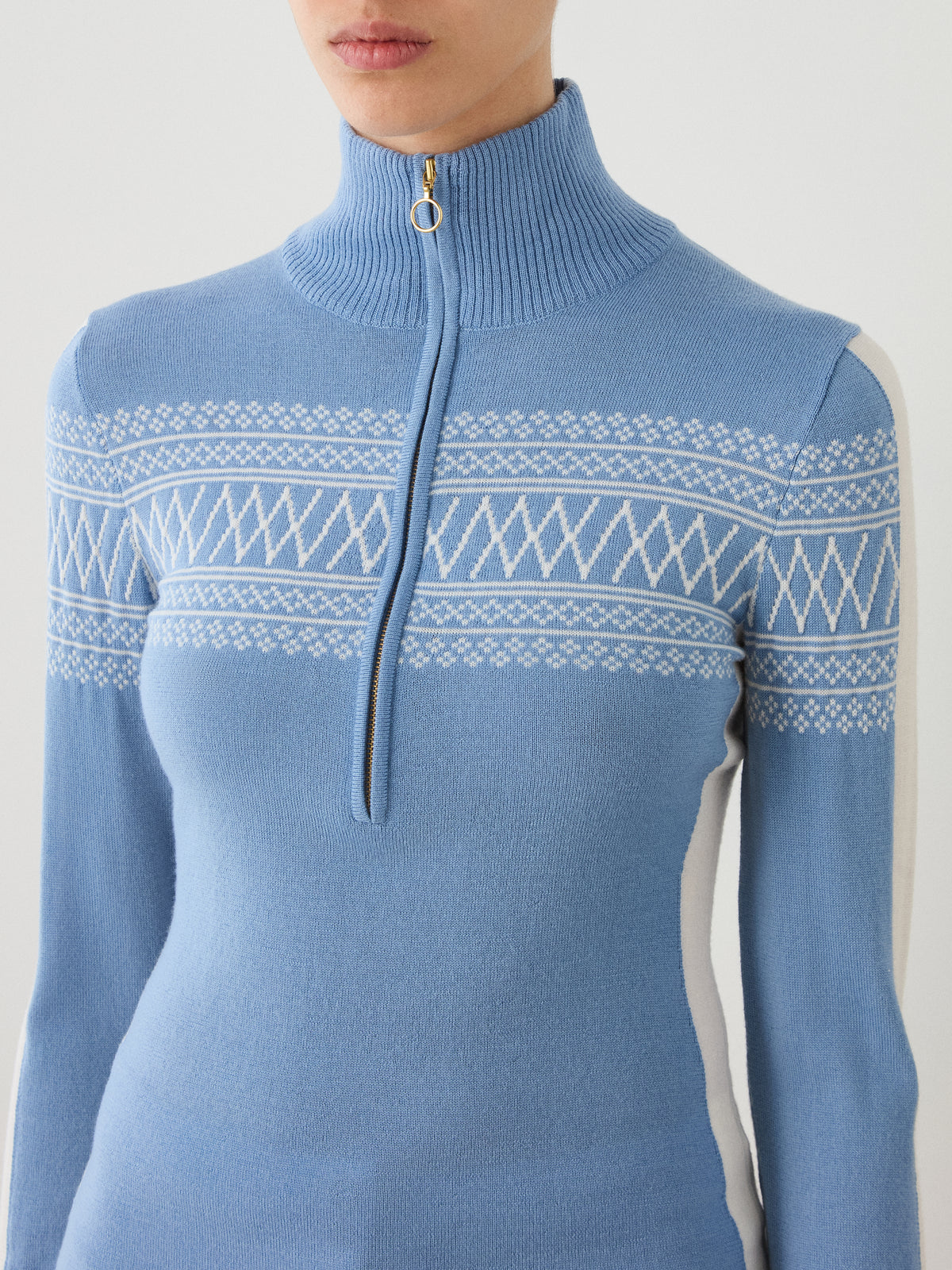 A person wearing the We Norwegians Signature 1/4-Zip Women sweater in blue, showcasing a white Setesdal pattern, a high ribbed collar, and the brand’s signature quarter-zip with a ring pull. Made from soft Merino wool, the image is cropped from shoulders to waist.