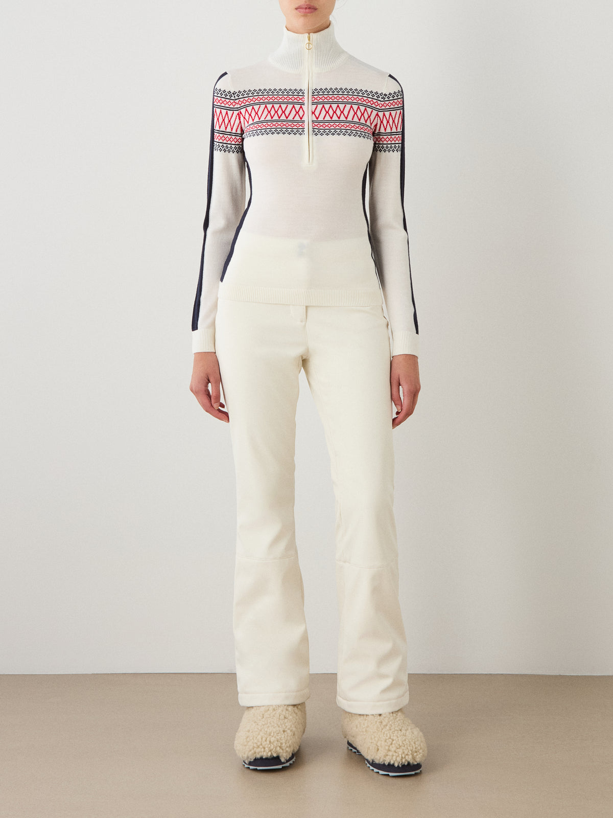 A person stands indoors facing forward against a plain background, dressed in white pants and fuzzy cream boots, and wearing the We Norwegians Signature 1/4-Zip Women’s sweater—a high-neck top featuring red and navy Setesdal patterns.