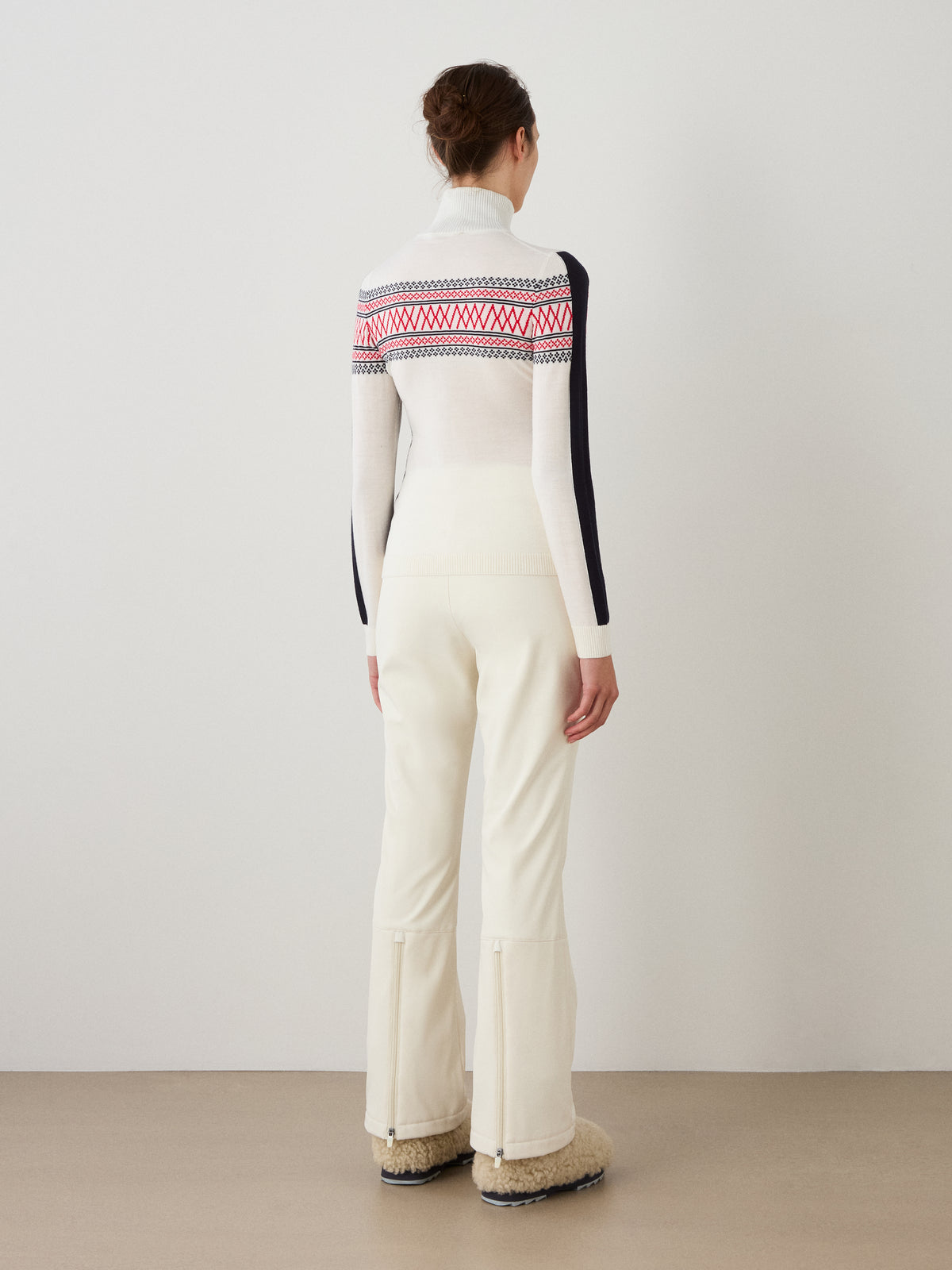 A woman stands indoors facing away, wearing We Norwegians’ Signature 1/4-Zip Women long-sleeve Merino Wool top in white with a subtle Setesdal pattern, paired with white pants and fuzzy beige boots against a plain wall and neutral floor.
