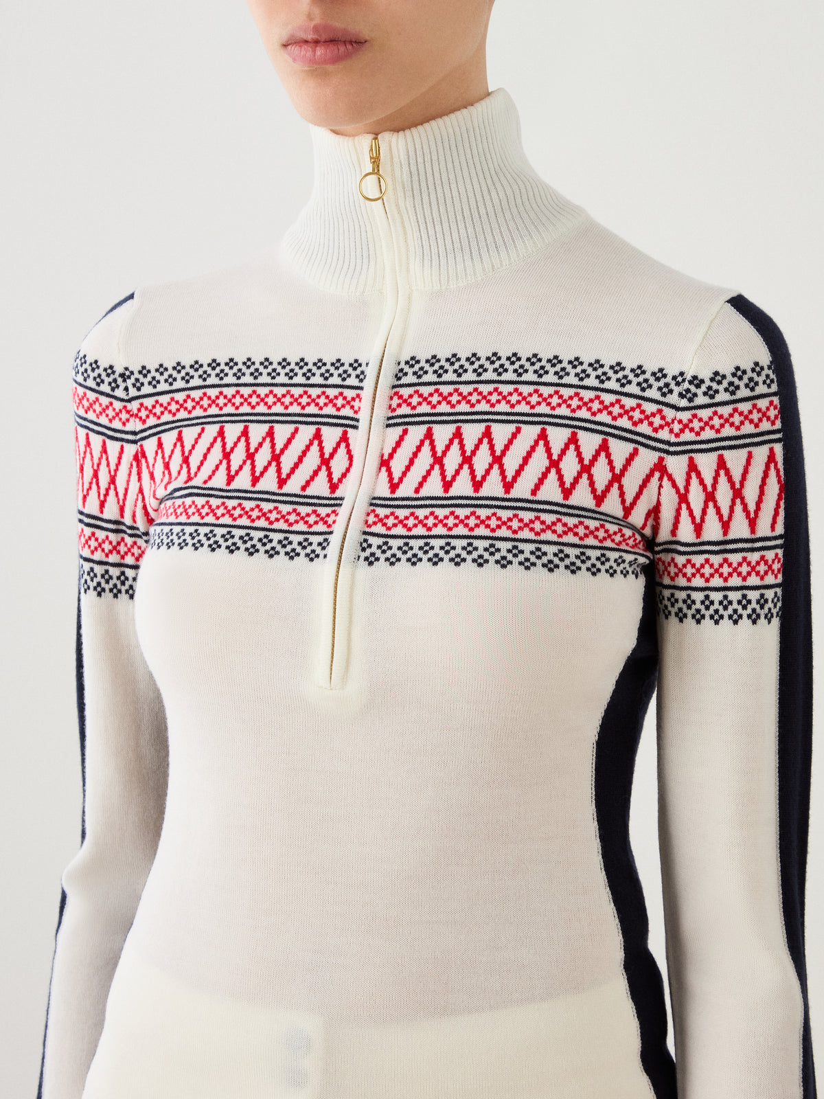 A person wearing the We Norwegians Signature 1/4-Zip Women sweater—a white Merino Wool turtleneck featuring a classic red and black Setesdal pattern across the chest, with a gold half-zip front—for timeless style and comfort.