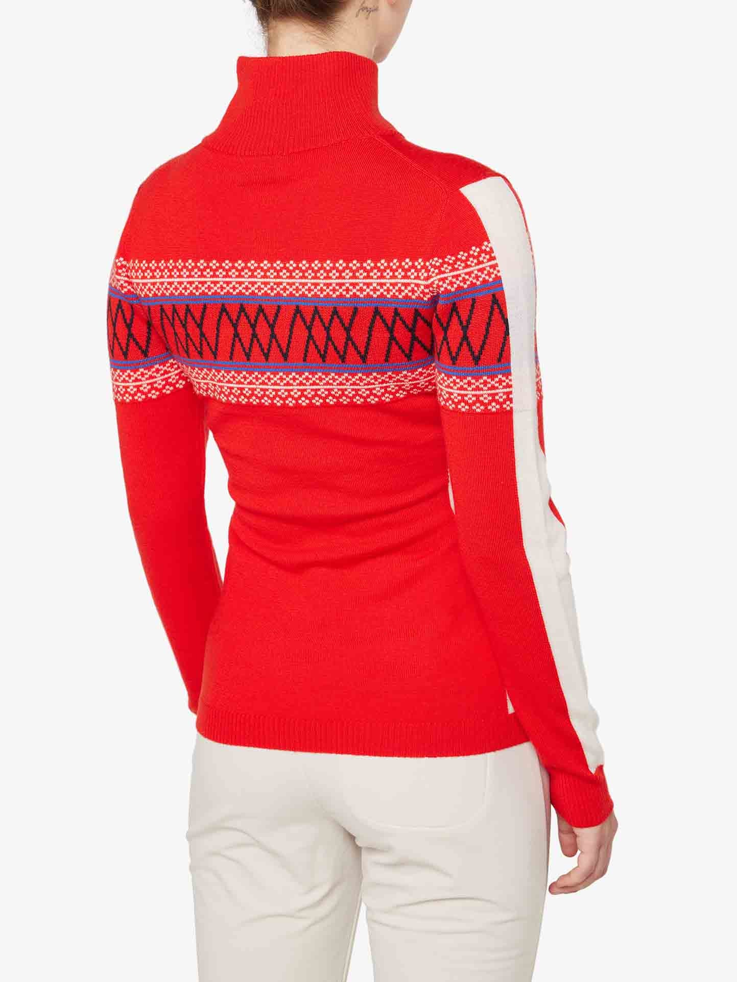 The person is wearing a Signature 1/4-Zip Women sweater by We Norwegians. The red Merino wool sweater features a Nordic Setesdal pattern with white and black geometric designs across the upper back and arms, complemented by white stripes running along the outer sleeves. They are also sporting white pants and are viewed from the back.