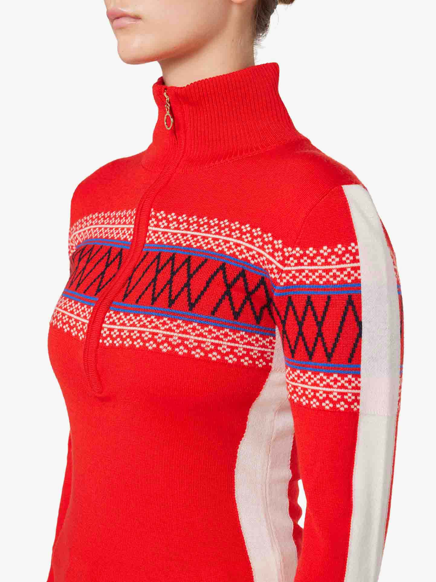 A person is shown from the neck down, wearing We Norwegians' Signature 1/4-Zip Women sweater in vibrant red. The fine Merino wool garment showcases intricate blue, white, and black geometric Setesdal patterns across the chest and sleeves, complemented by white panels on the sides. The collar is ribbed and zipped up.