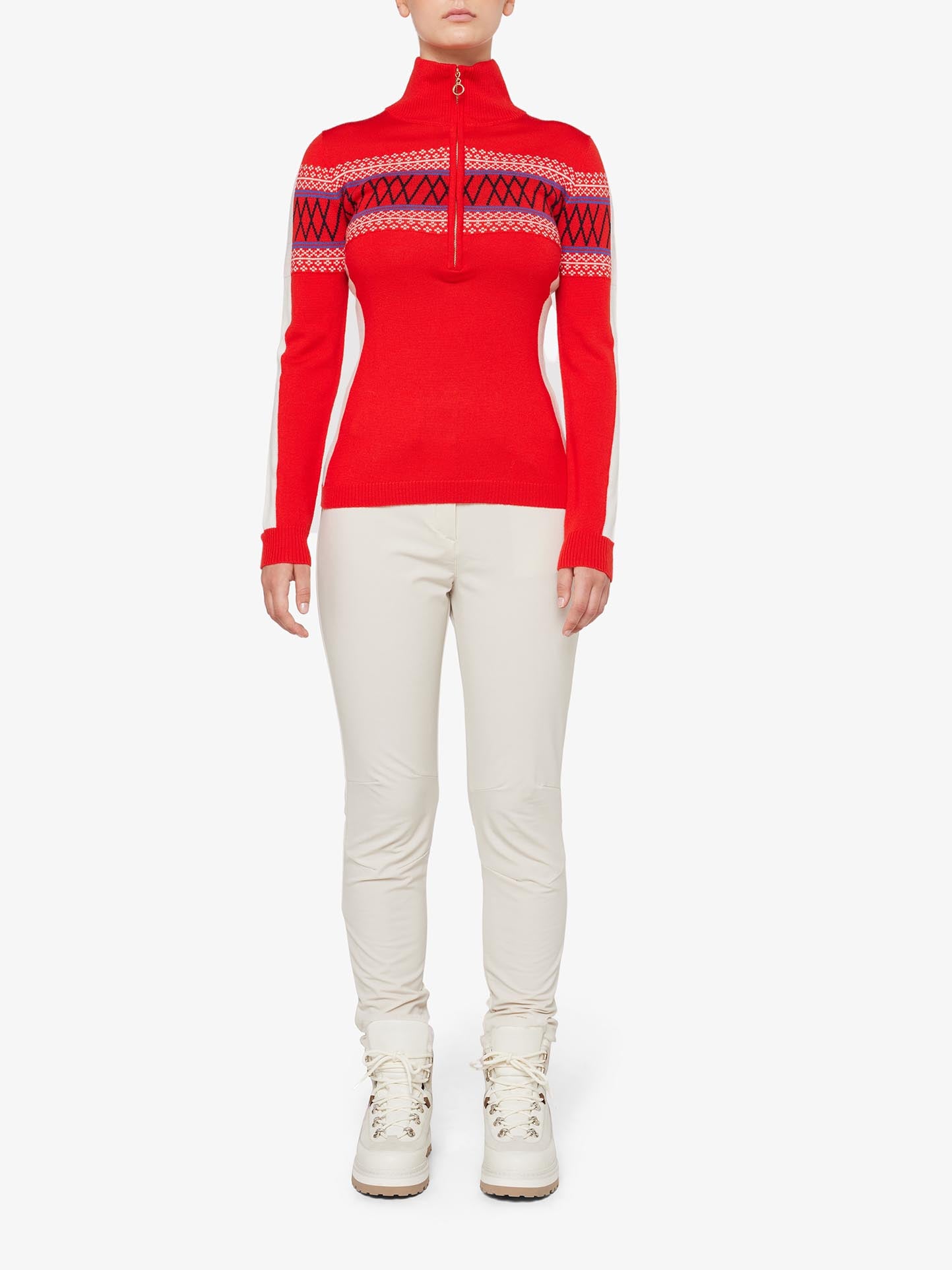 A person is wearing the We Norwegians Signature 1/4-Zip Women in bright red, crafted from fine Merino wool and featuring a traditional Setesdal pattern around the chest and upper arms. They have paired it with white fitted pants and lace-up boots. The outfit appears to be suitable for winter or skiing, with the person standing against a plain white background.