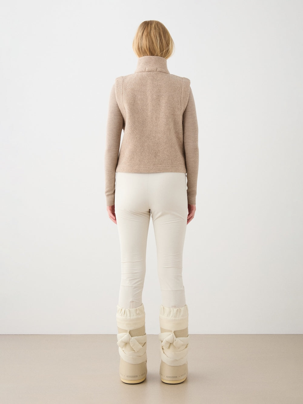 A person stands facing away, wearing the We Norwegians Alta Vest Women—a felted merino wool layering essential—over a beige turtleneck sweater, paired with cream pants and off-white winter boots. The plain, minimalistic background highlights the vest’s versatile style for colder months.