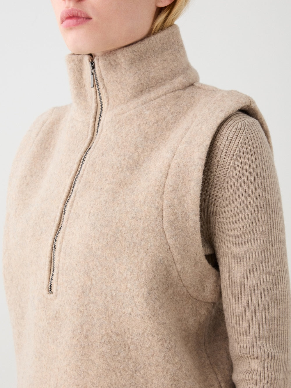 A person wearing the We Norwegians Alta Vest Women—a beige, sleeveless, high-collar zip vest perfect for layering—over a ribbed long-sleeve top, shown from the shoulders up against a plain background.