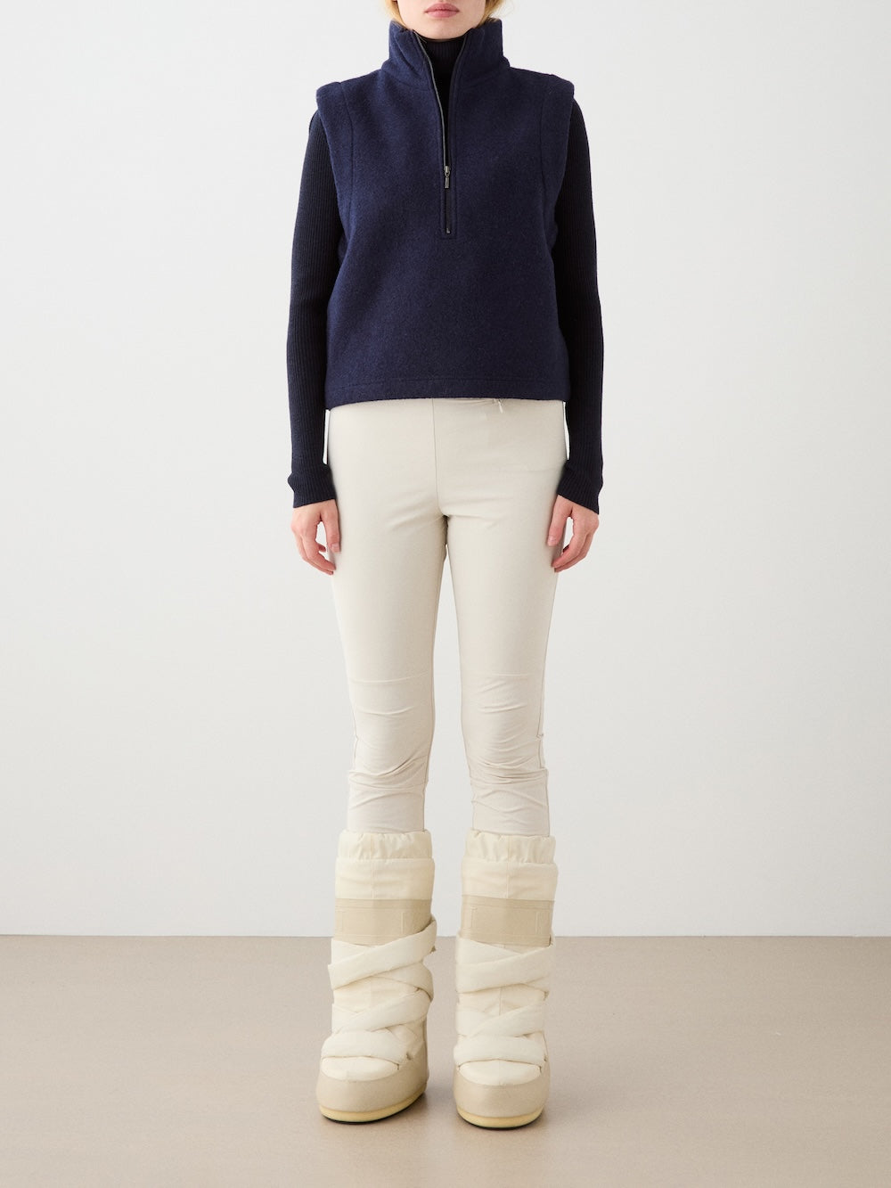 A person wearing the We Norwegians Alta Vest Women in navy merino wool over a black long-sleeve top, cream fitted pants, and chunky cream snow boots stands against a plain background. Their face is not visible.