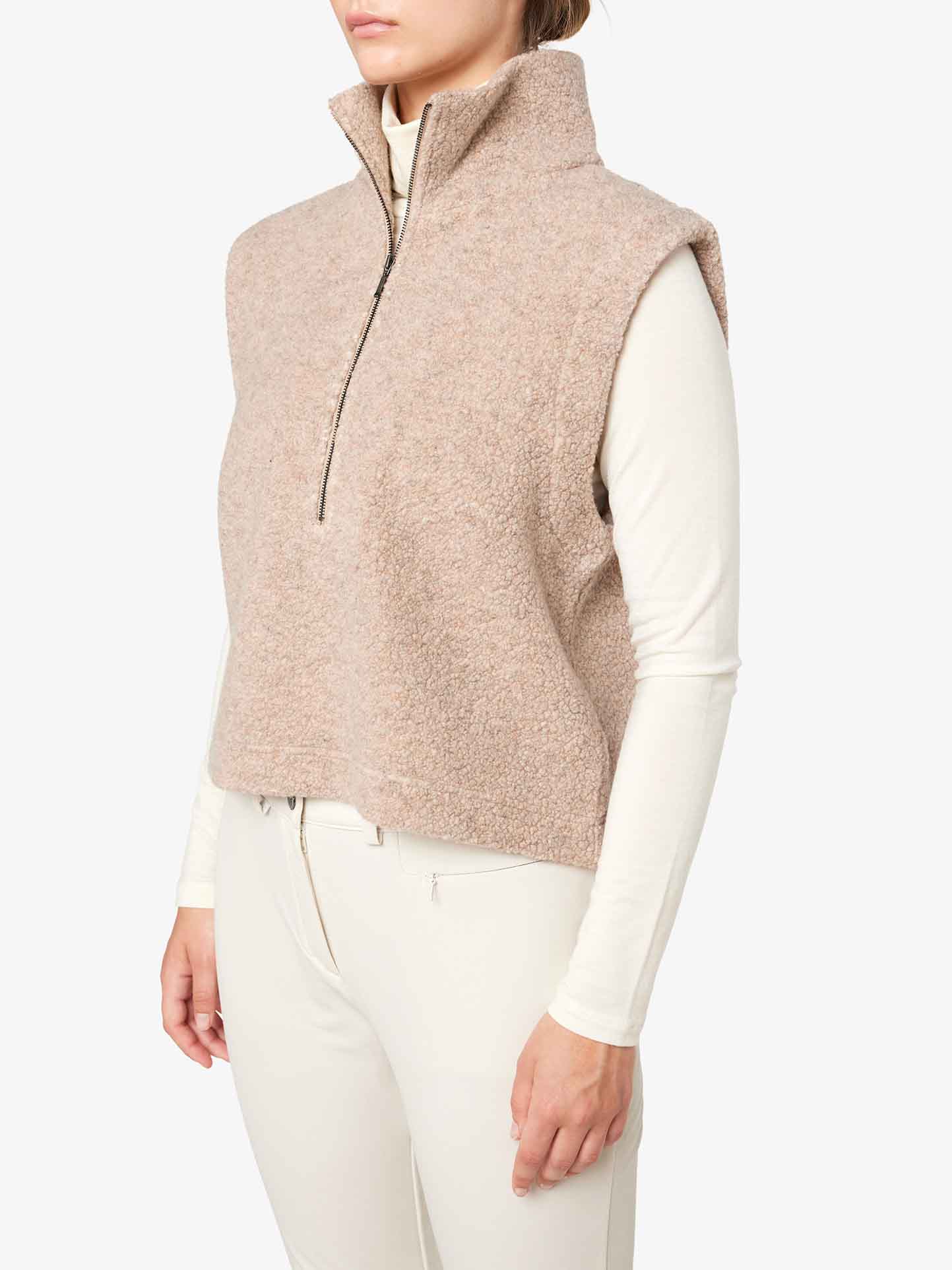 A person is layering an Alta Vest Women by We Norwegians, featuring a high collar and a partial front zipper, over a white long-sleeve top. The outfit is paired with white pants against a plain, light-colored background. The person's face is not visible in the image.