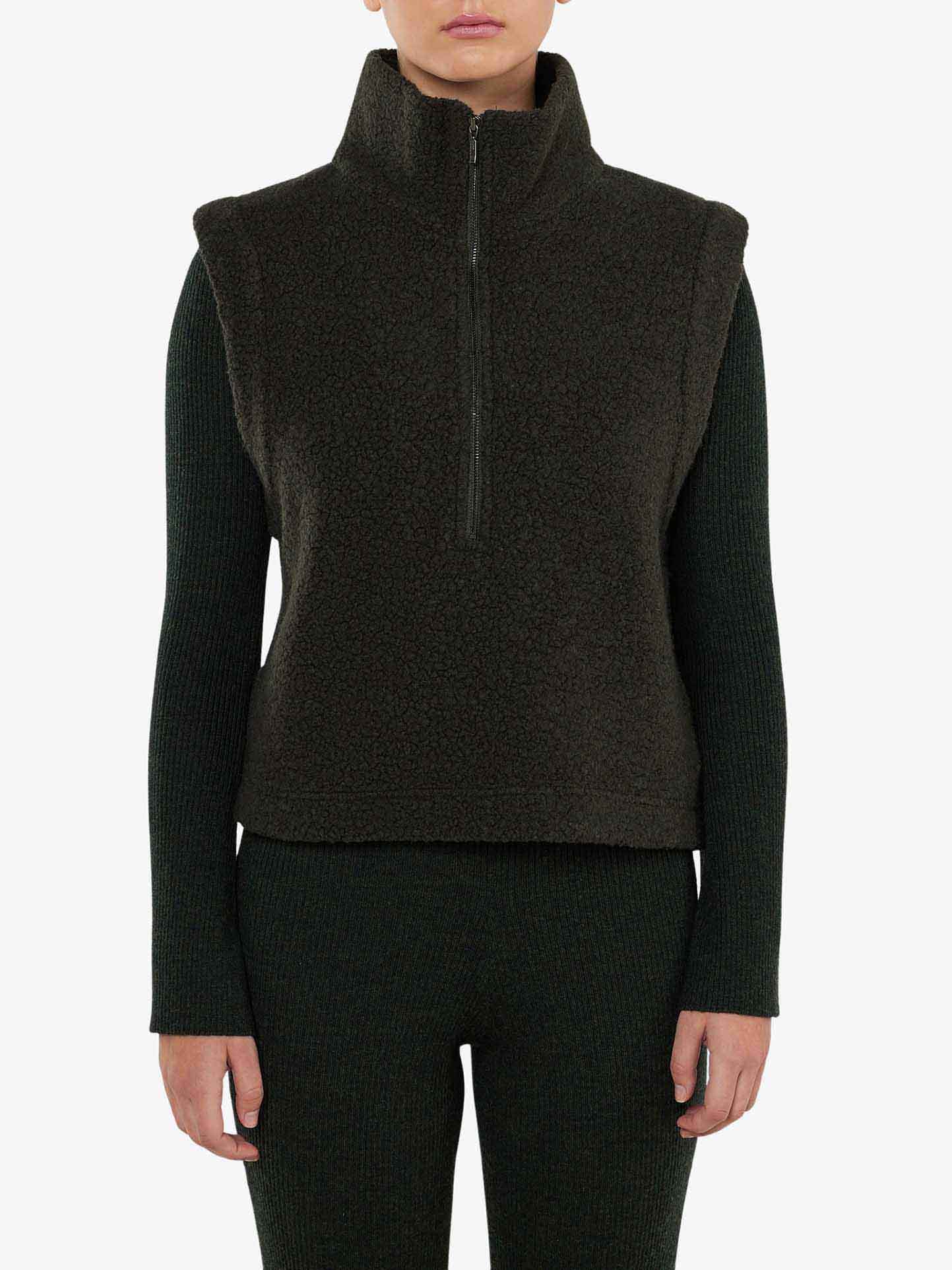 A person is wearing the dark green, textured Alta Vest Women by We Norwegians, featuring a high collar and half-zip front, over a ribbed long-sleeve sweater and matching pants. The Merino wool vest provides excellent layering options, giving the outfit a cozy, winter-ready appearance.