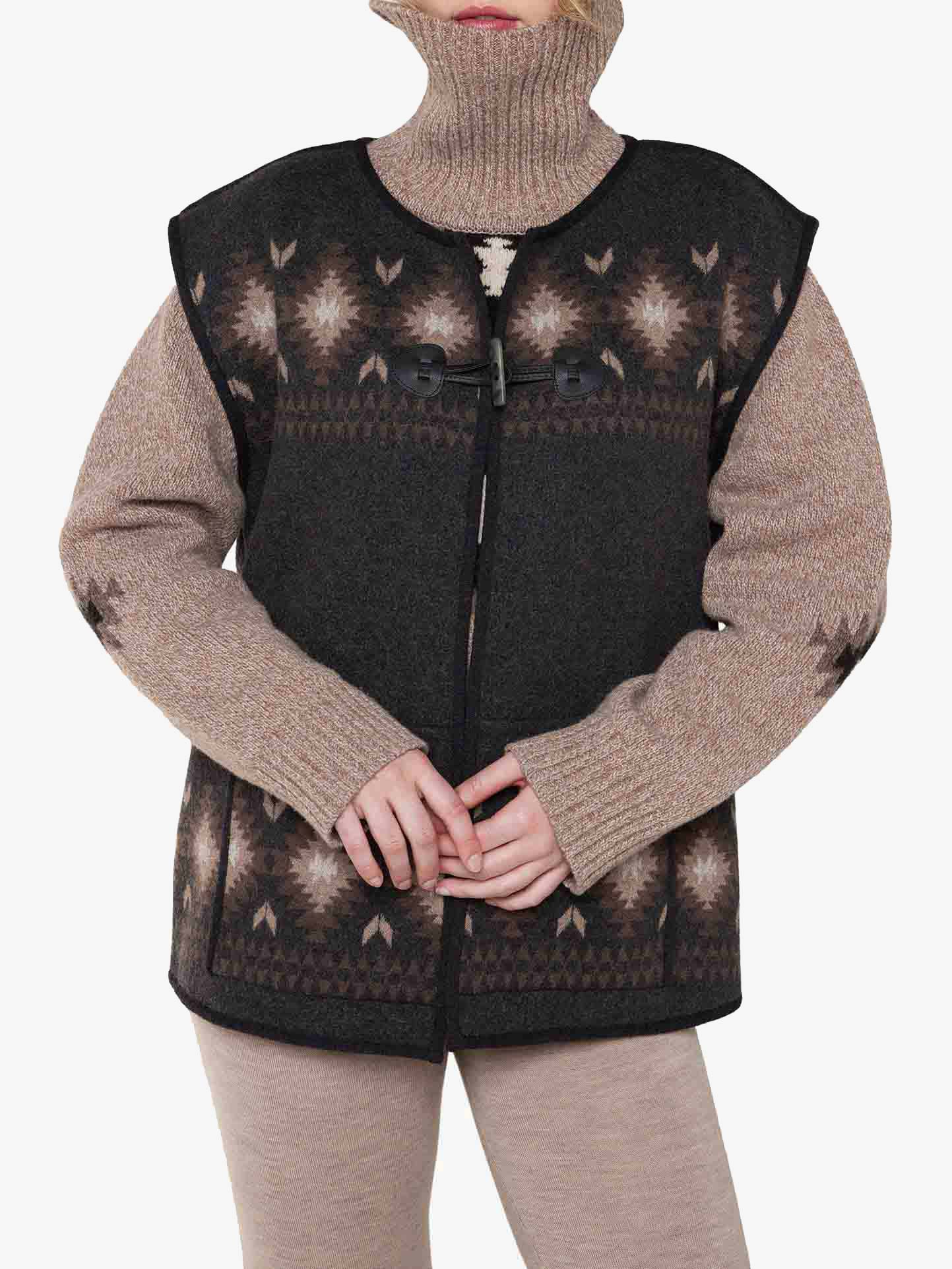 A person wearing a high-neck beige sweater underneath the *Hovden Vest Women* by *We Norwegians*, a dark, sleeveless knitted vest featuring geometric designs inspired by Sámi culture. The vest, possibly made from merino wool, includes a front clasp closure. They are also dressed in beige pants, and their hands are clasped together in front of them against a plain white background.