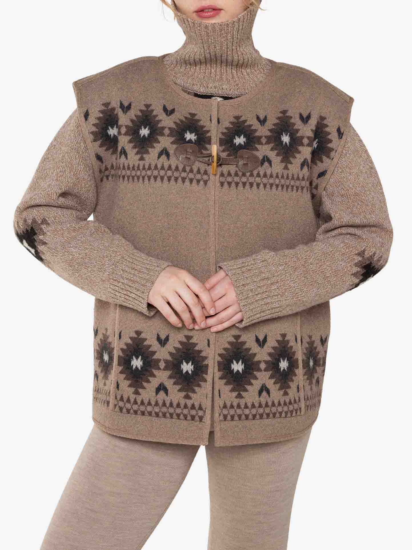 A person wearing the Hovden Vest Women by We Norwegians, a neutral-toned, patterned turtleneck sweater and a boiled wool cardigan with geometric and floral designs in shades of brown, black, and white. The cardigan features a toggle closure at the neck. The person is standing against a plain white background.