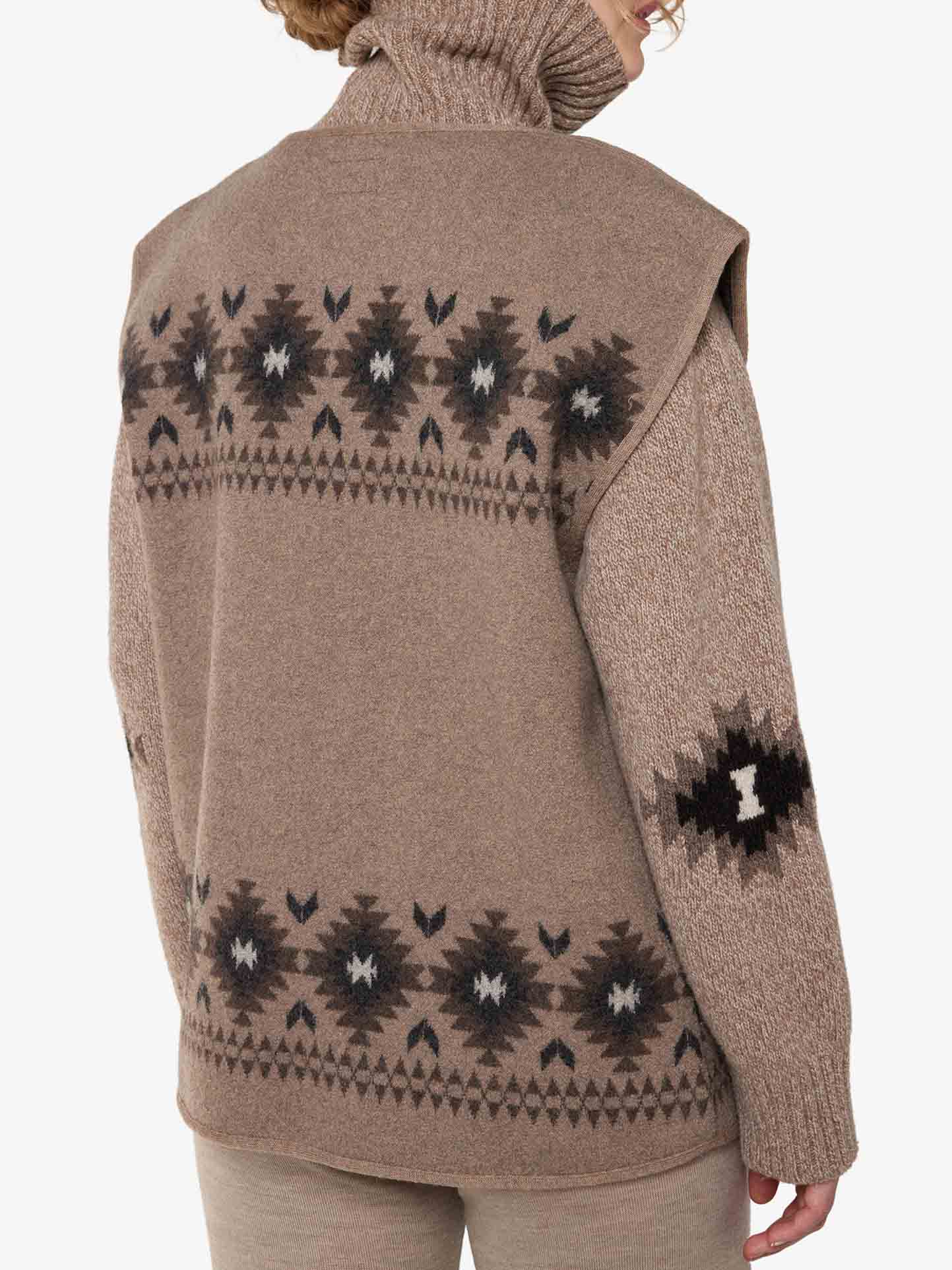 A woman sporting the Hovden Vest Women by We Norwegians, a beige turtleneck sweater with intricate black and brown geometric patterns on the back and sleeves, reminiscent of Sámi Culture Design. The soft, knitted texture adds to its cozy and warm appearance. She has short hair and is turned away from the camera.