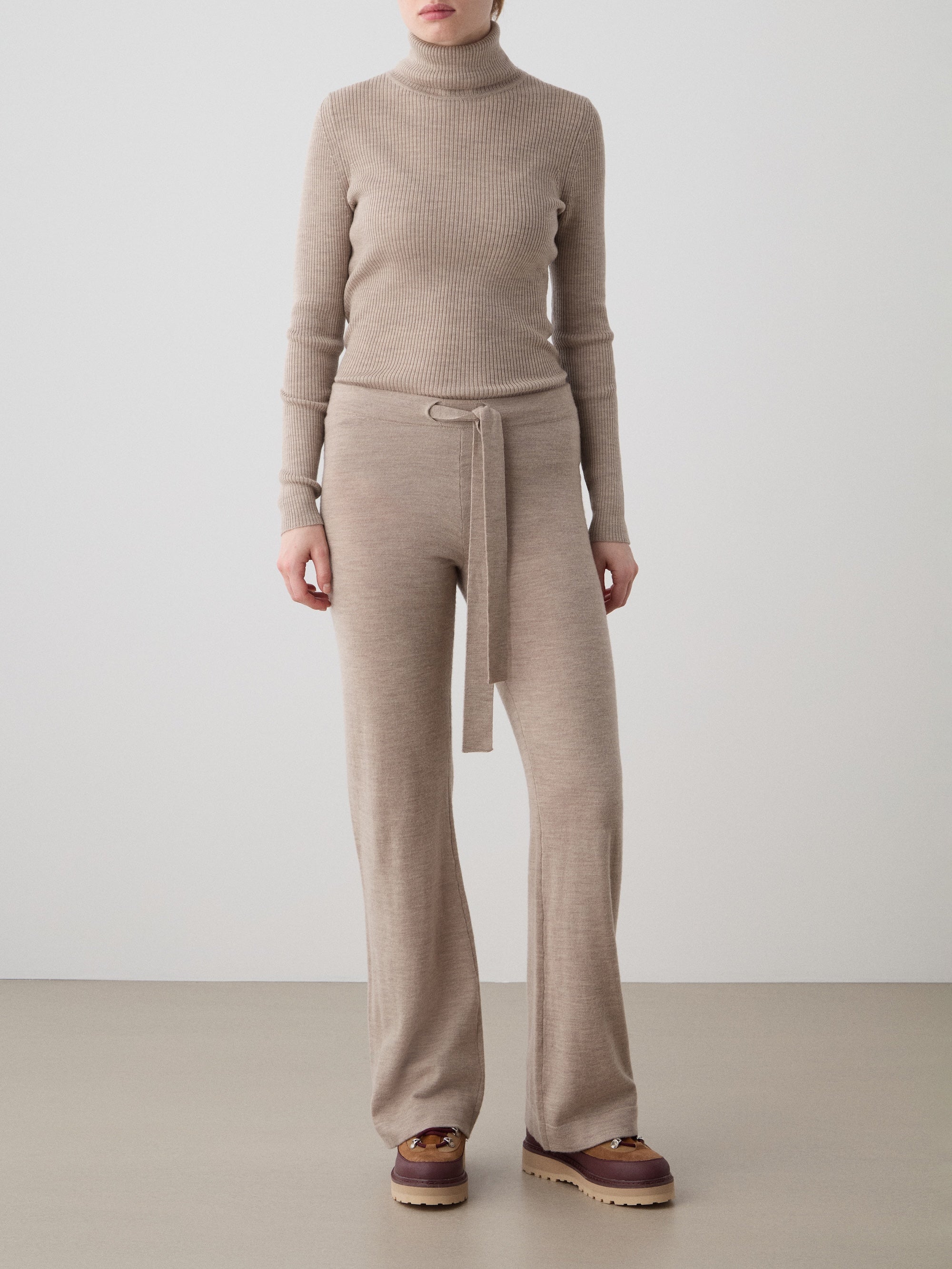 A person wearing We Norwegians’ Geilo Belted Pant Women in beige with a matching turtleneck and brown platform shoes, standing against a plain background.