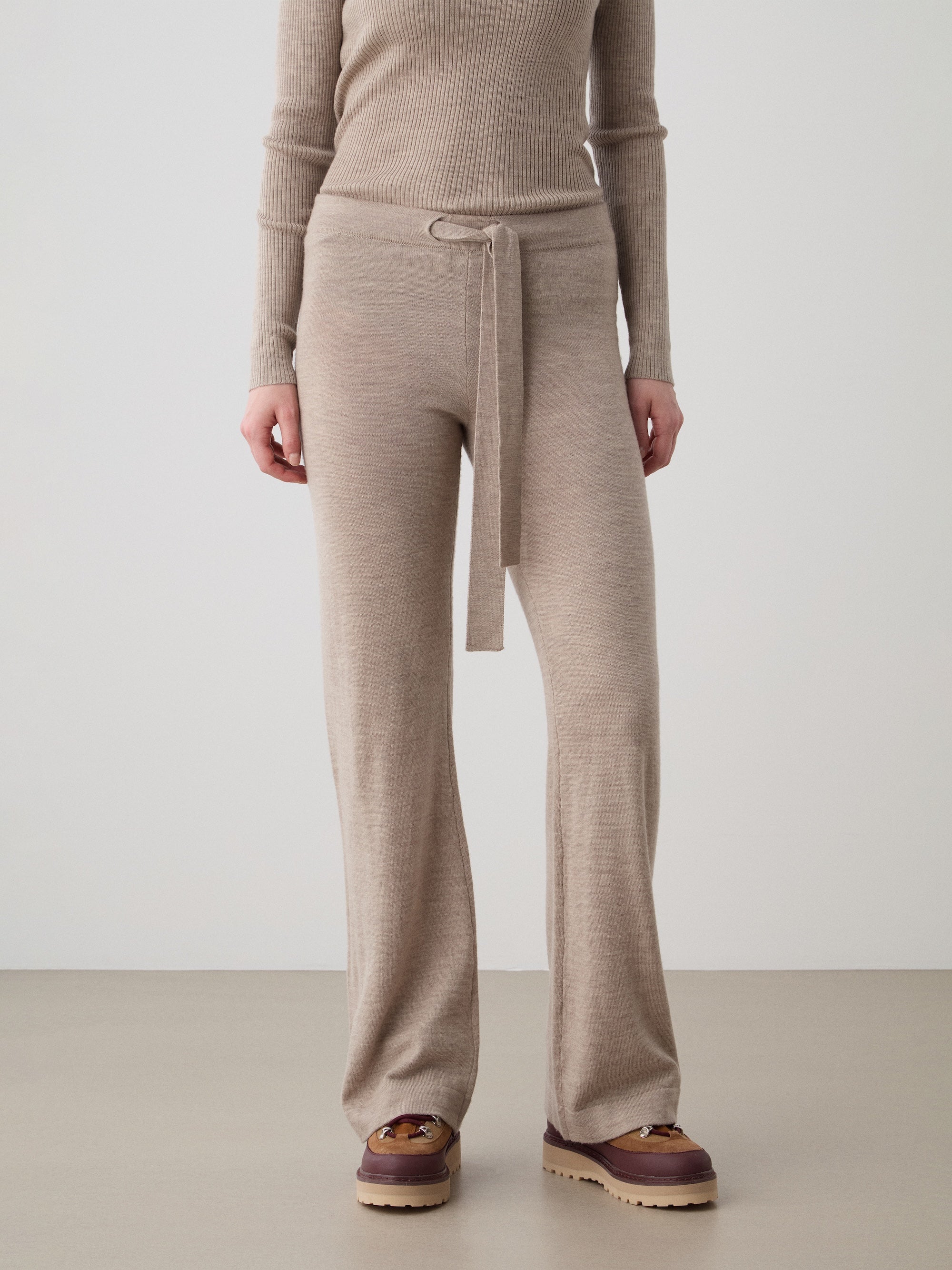 A person wears the We Norwegians Geilo Belted Pant for women with a beige ribbed long-sleeve top and brown shoes, standing against a plain background.