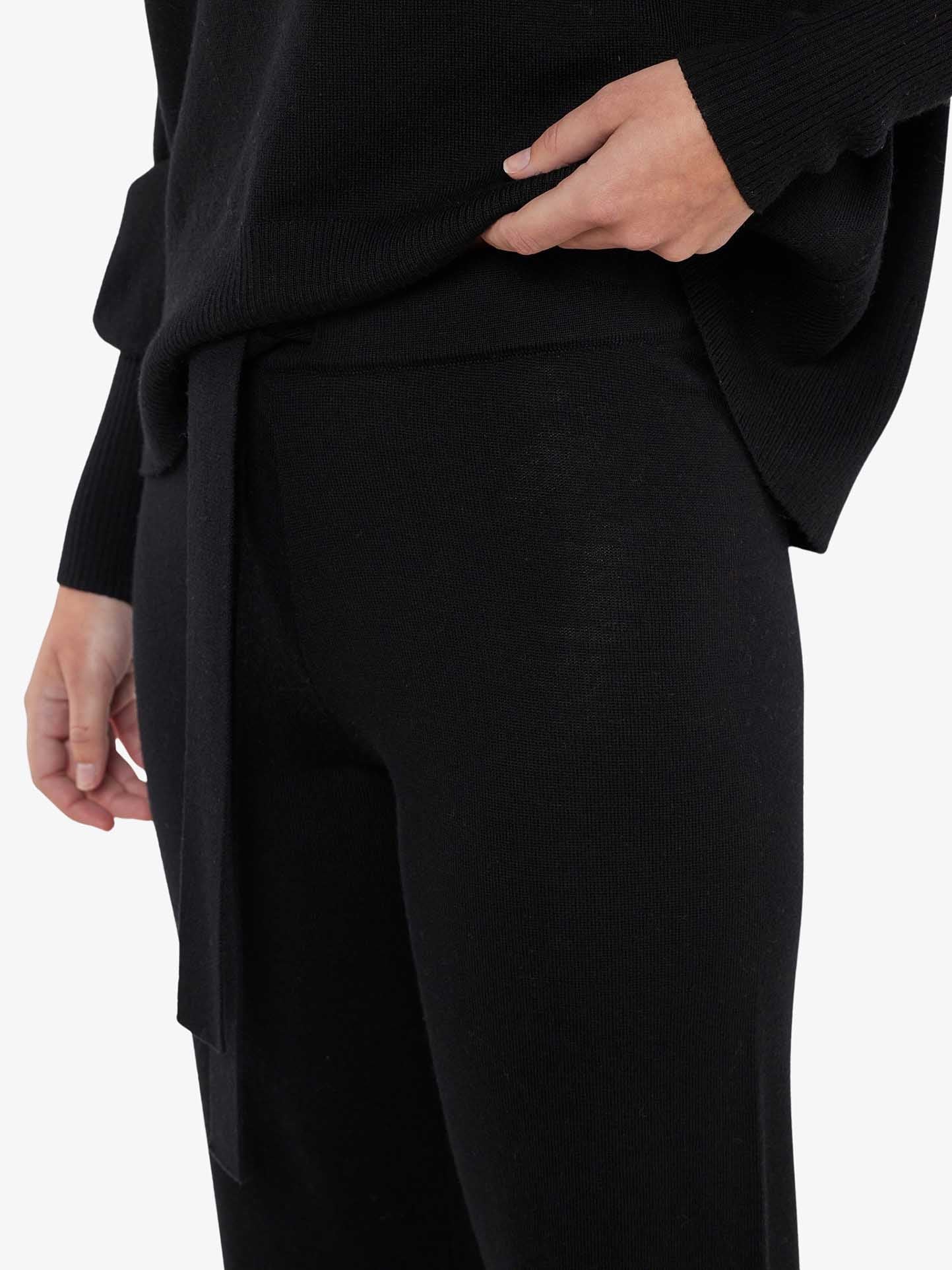 A woman is shown from her shoulders to her thighs, wearing a black long-sleeve top and comfortable Geilo Belted Pant Women from We Norwegians, featuring a tie waist. Her left hand is partially tucked into her pocket, and her right arm hangs down by her side. The Merino pants, made from 100% Merino Wool, stand out against the white background.