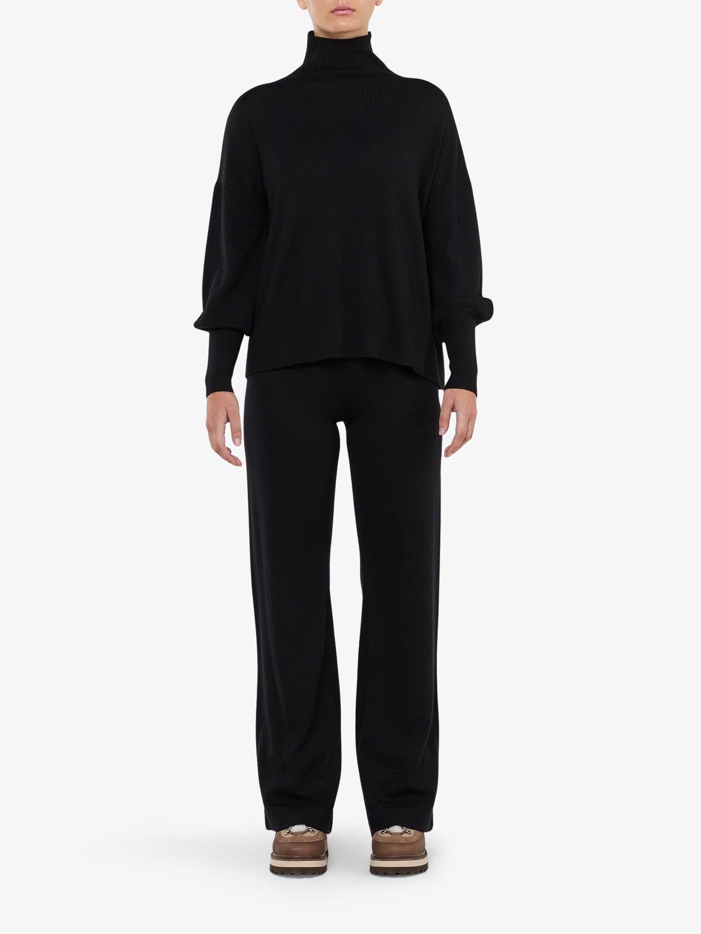 A person faces forward wearing a loose black turtleneck, We Norwegians Geilo Belted Pant Women in black, and brown shoes against a plain white background.
