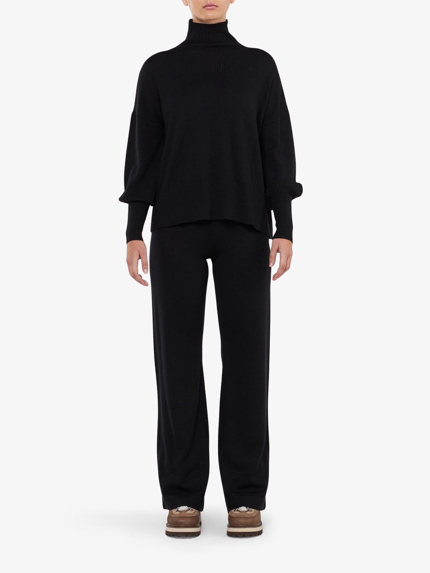 A person is standing against a plain background, wearing a black long-sleeve turtleneck sweater paired with the Geilo Belted Pant Women from We Norwegians. The individual has brown shoes on and has their arms relaxed by their sides. The comfortable soft pants, made of 100% Merino Wool, add a touch of luxury to the minimalist ensemble.