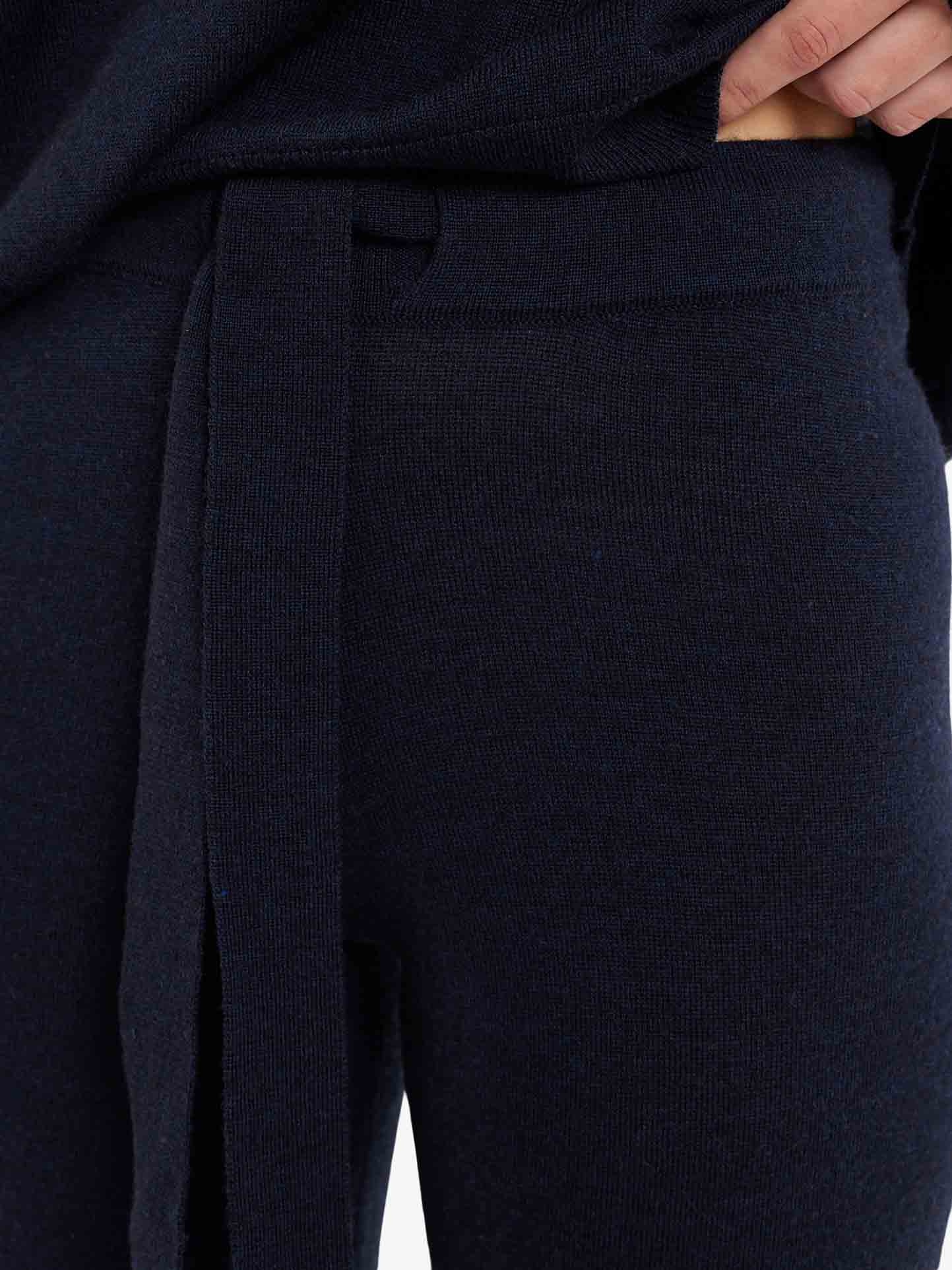 Close-up of a person wearing the Geilo Belted Pant Women by We Norwegians, dark-colored and knitted with a matching tie belt. The person's hand is partially visible, resting near the waistband. The fabric appears soft and comfortable, making these 100% Merino Wool pants perfect for casual or lounge wear.