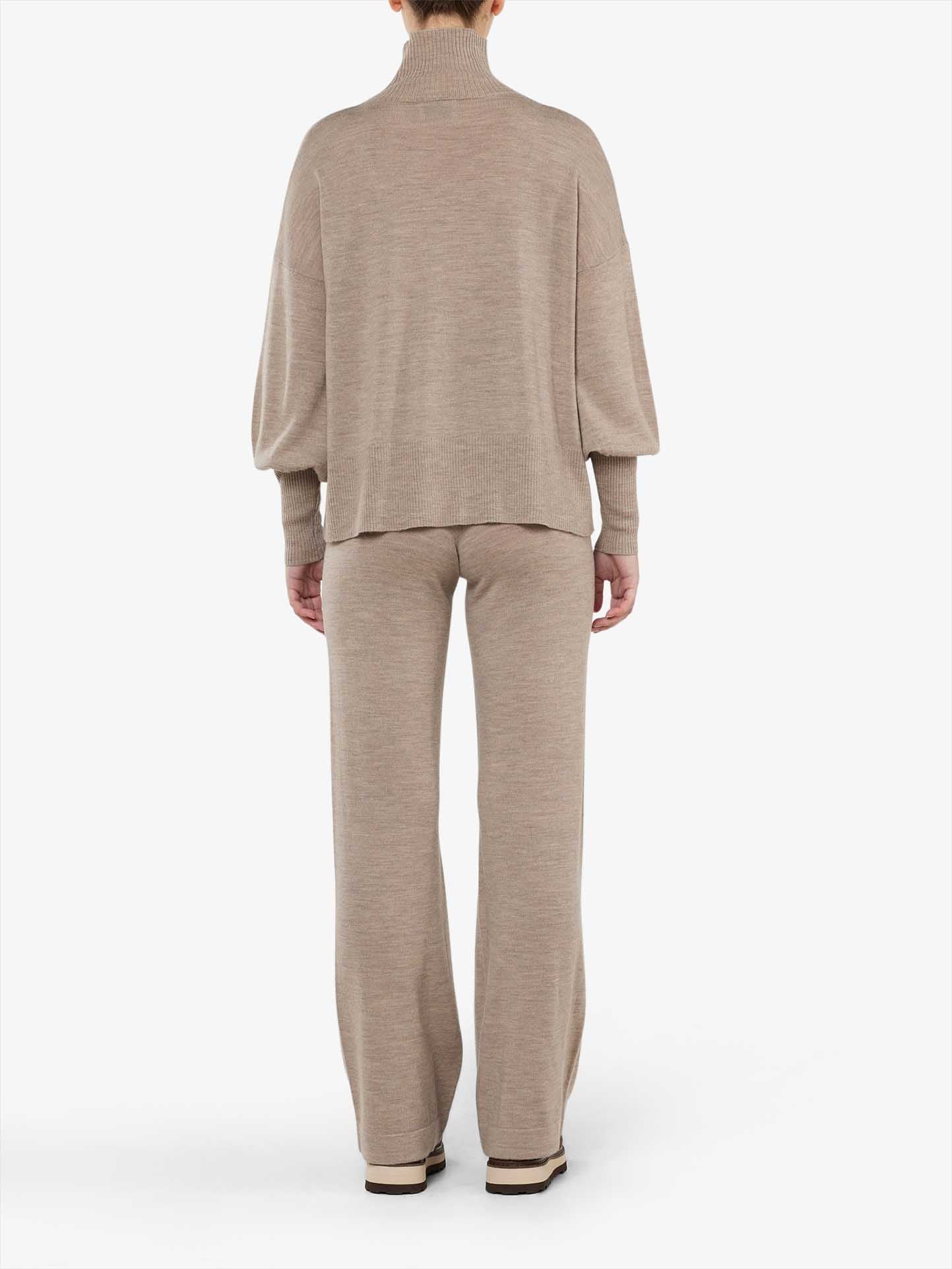 A person is standing facing away from the camera, wearing a beige, long-sleeved turtleneck sweater paired with matching Geilo Belted Pant Women by We Norwegians. The outfit appears to be made of a comfortable 100% Merino Wool knit material. The background is plain white.