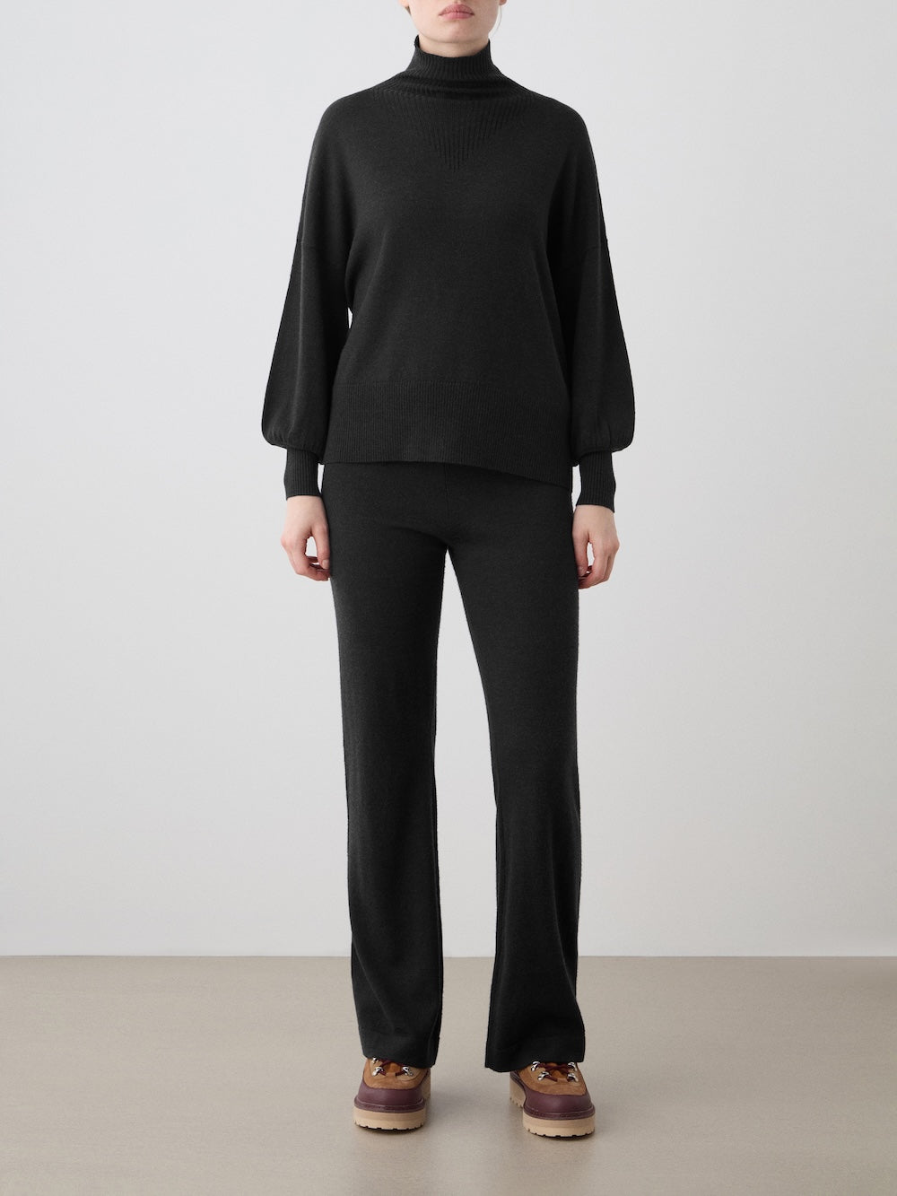 A woman stands against a plain light background, wearing the We Norwegians Geilo Oversized Sweater in black with matching black pants and brown platform shoes. Her arms are relaxed by her sides.