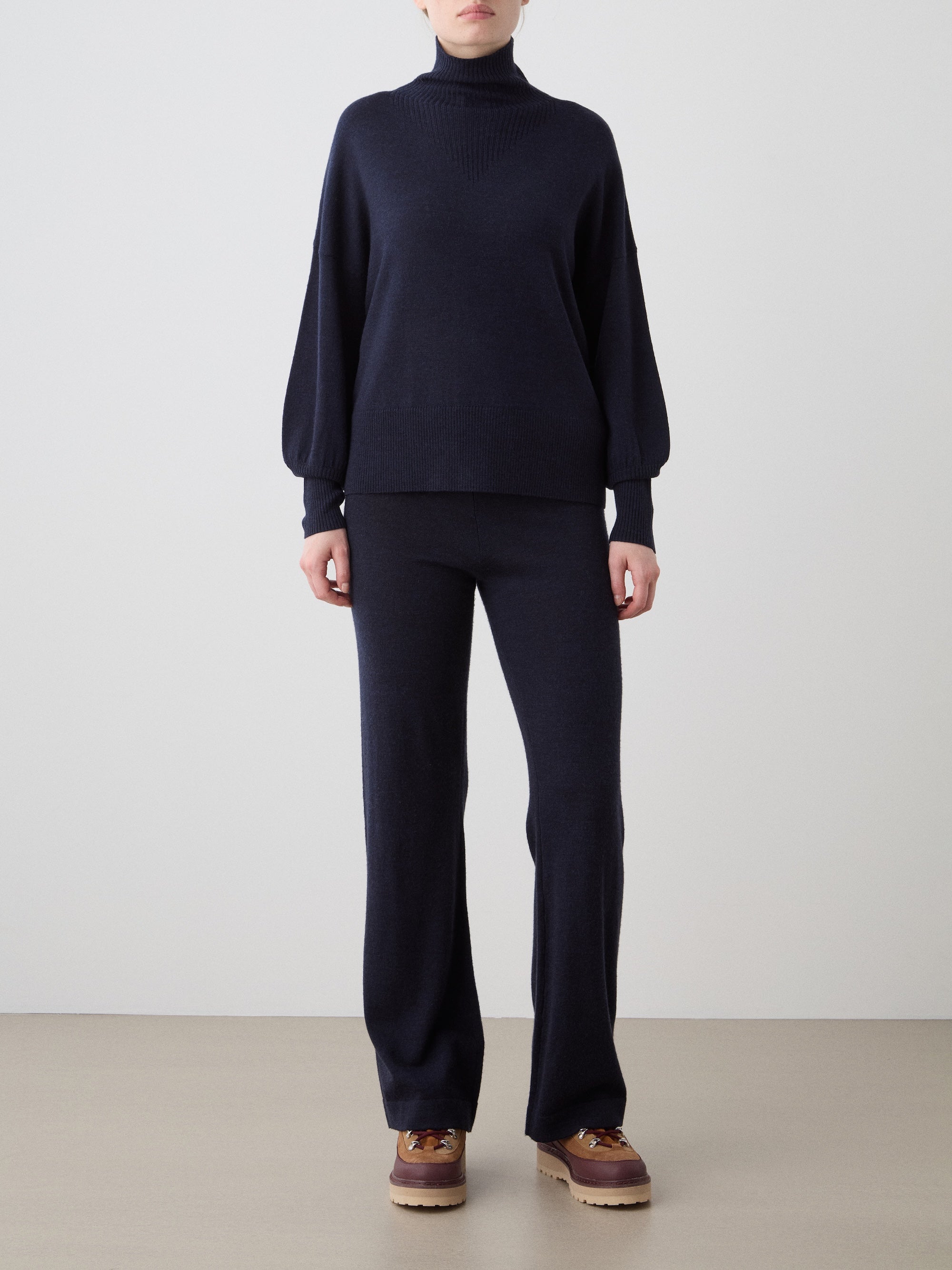 A woman wears the We Norwegians Geilo Oversized Sweater in dark navy with matching pants and brown lace-up shoes, standing against a plain light background.
