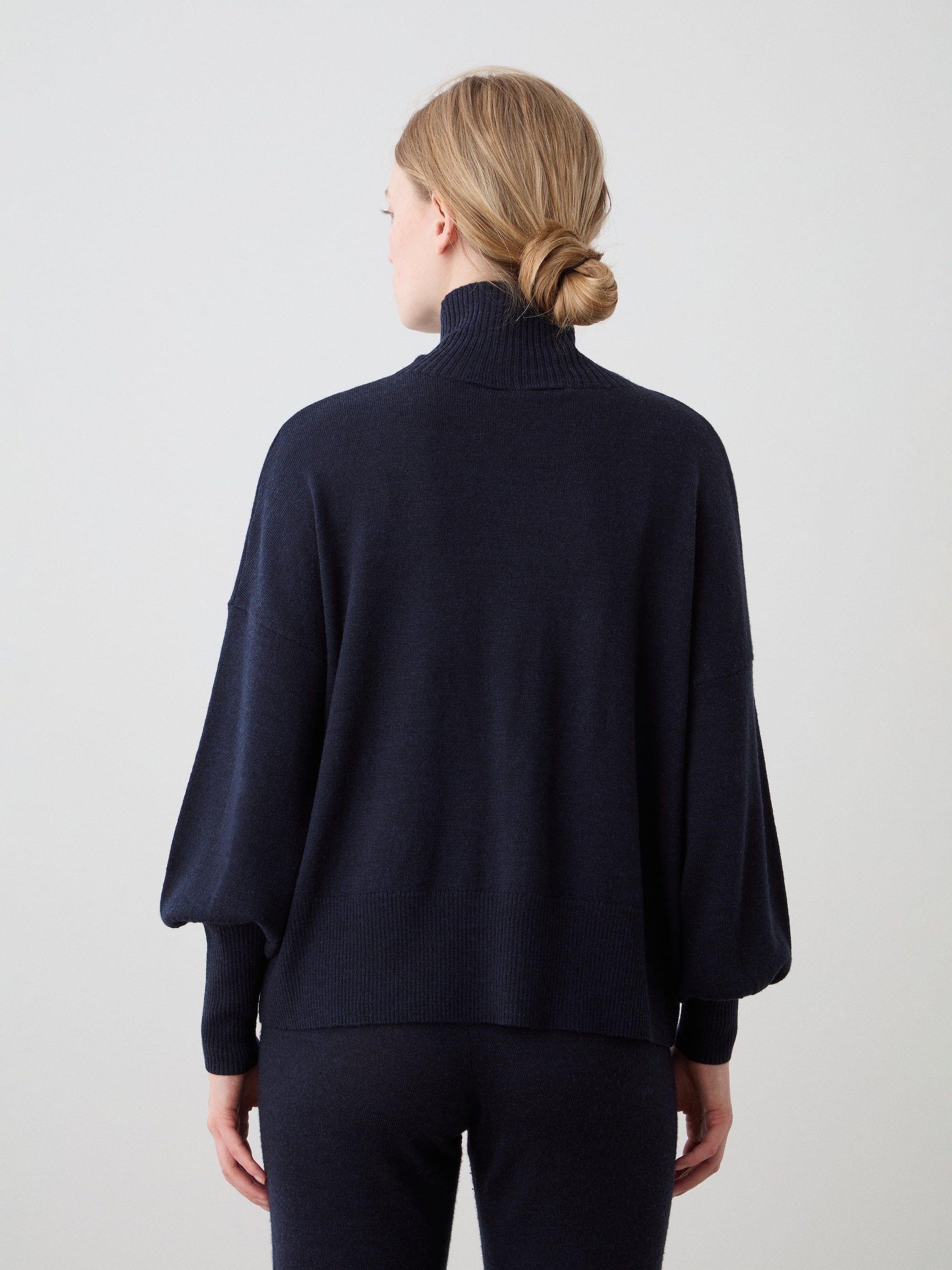 A woman with blonde hair in a low bun stands with her back to the camera, wearing the We Norwegians Geilo Oversized Sweater for women in dark navy and matching pants, set against a plain light background.