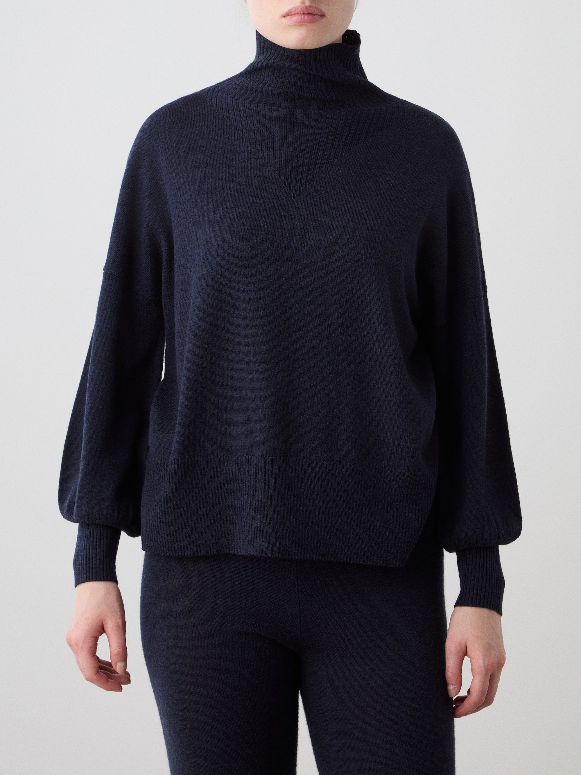 A woman models the We Norwegians Geilo Oversized Sweater in dark navy merino wool with long sleeves and matching pants, standing against a plain light background. The relaxed fit and partially out-of-frame face create an effortless look.