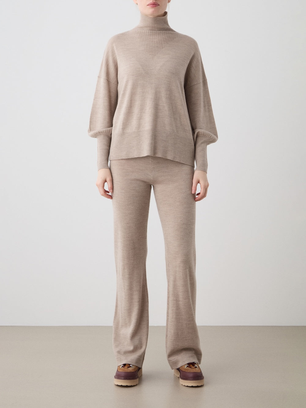 A person stands against a plain background wearing the We Norwegians Geilo Oversized Sweater for women, paired with matching wide-leg pants and brown slip-on shoes, with their arms relaxed by their sides.