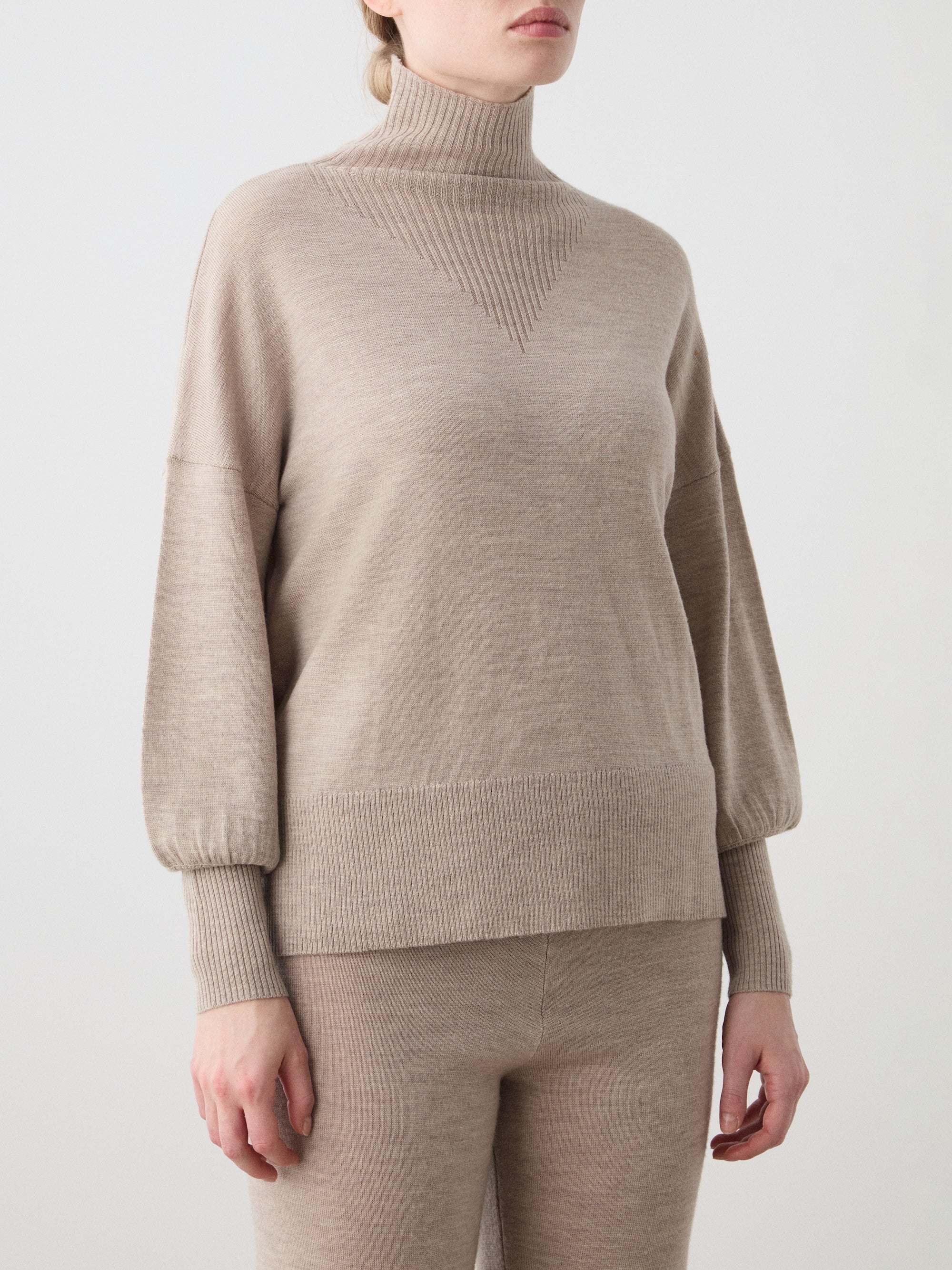 A woman wears the Geilo Oversized Sweater by We Norwegians—an elegant, lightweight beige knit turtleneck with long sleeves—paired with matching pants, standing against a plain light background.