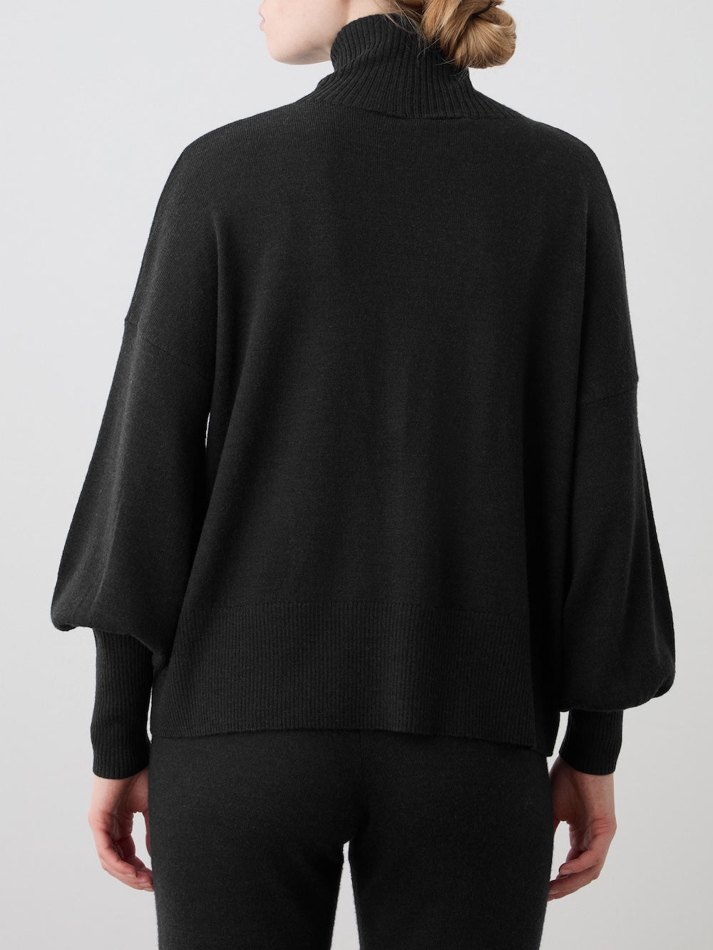 A person with blond hair in a low bun is seen from behind, wearing the We Norwegians Geilo Oversized Sweater for women with matching black pants, styled as a chic lounge set against a plain light background.