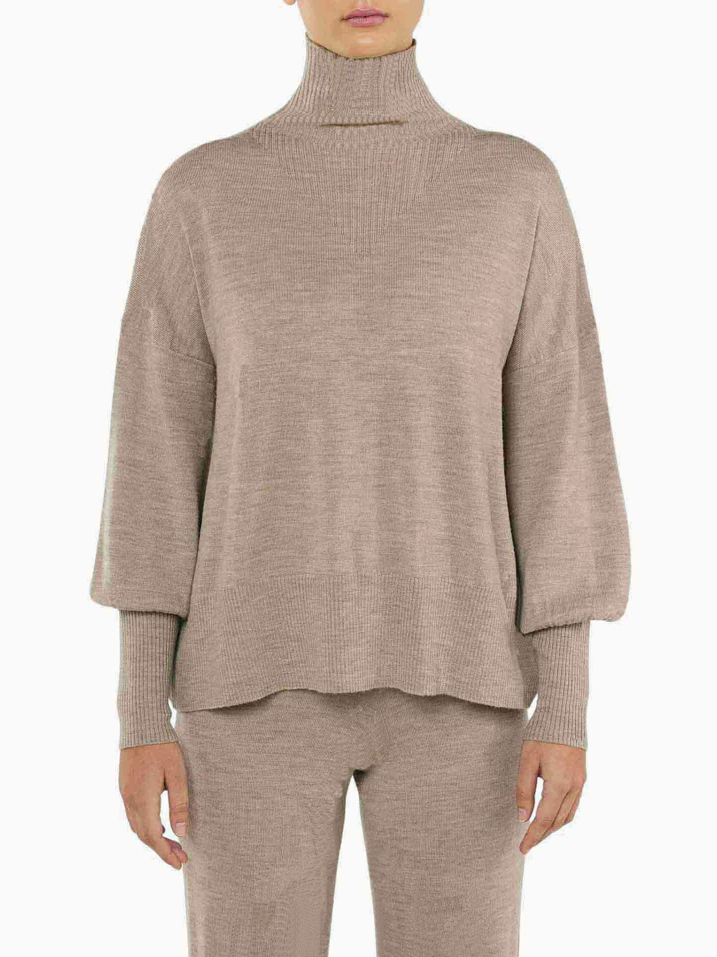 A person dressed in the Geilo Oversized Sweater Women by We Norwegians, featuring a loose-fitting design, long sleeves, a high turtleneck, and ribbed cuffs in beige merino wool. The ensemble is completed with matching beige knitted pants. The background is white, and the person's head is not visible in the frame.