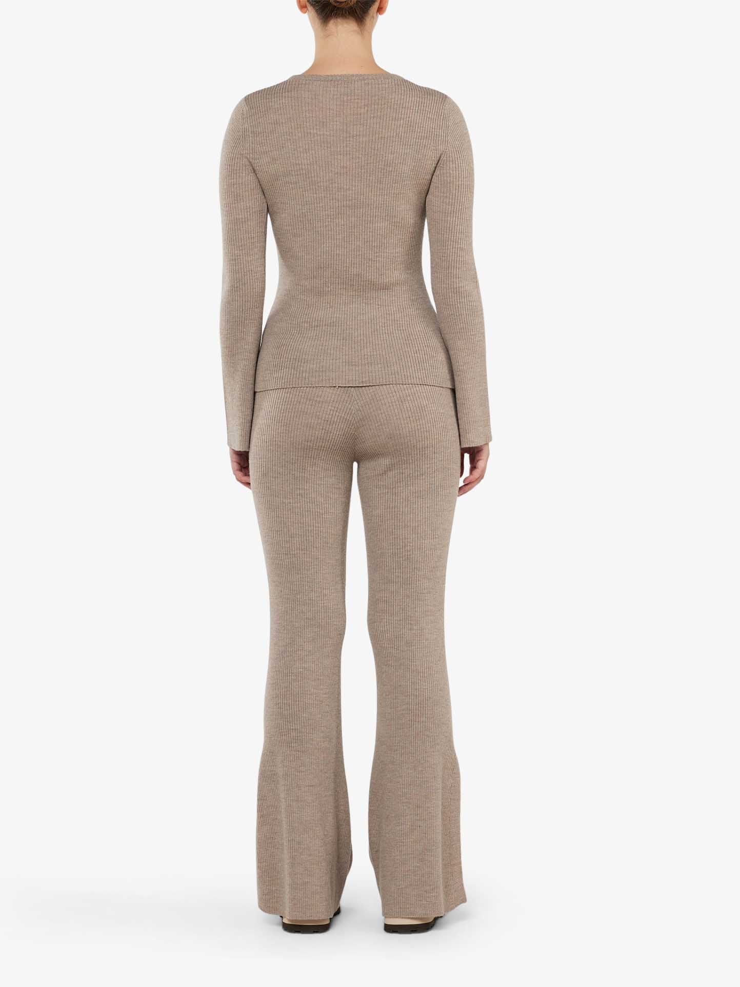 A person wearing the Geilo Ribbed Pant Women by We Norwegians, paired with a beige ribbed knit sweater, stands against a plain white background, facing away from the camera.